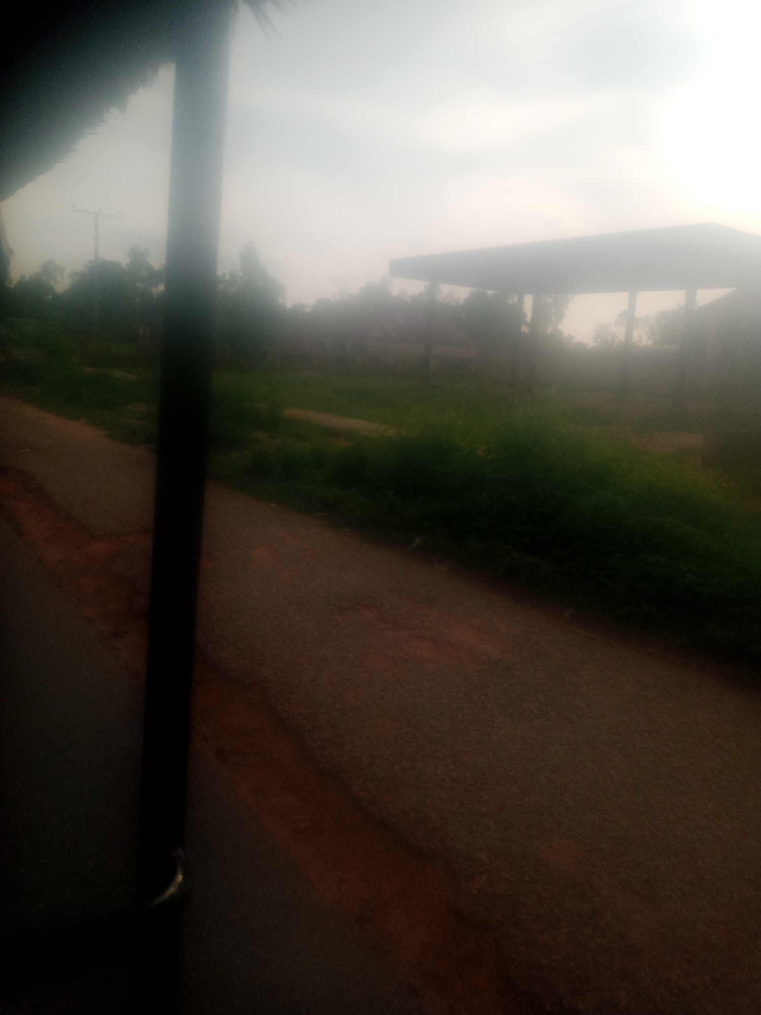 The image depicts a blurred outdoor scene, likely taken from a moving vehicle. A dark, vertical pole or structure dominates the left side of the frame. In the background, a light-colored, possibly gas station canopy, is visible through a hazy atmosphere, suggesting overcast conditions or dust. Trees and vegetation form a green band across the middle ground. A road or paved surface is present in the foreground, with a reddish-brown dirt shoulder. The overall impression is one of low visibility and ambient light. The location in Jauro Yinu, Nigeria, is inferred from the provided context.