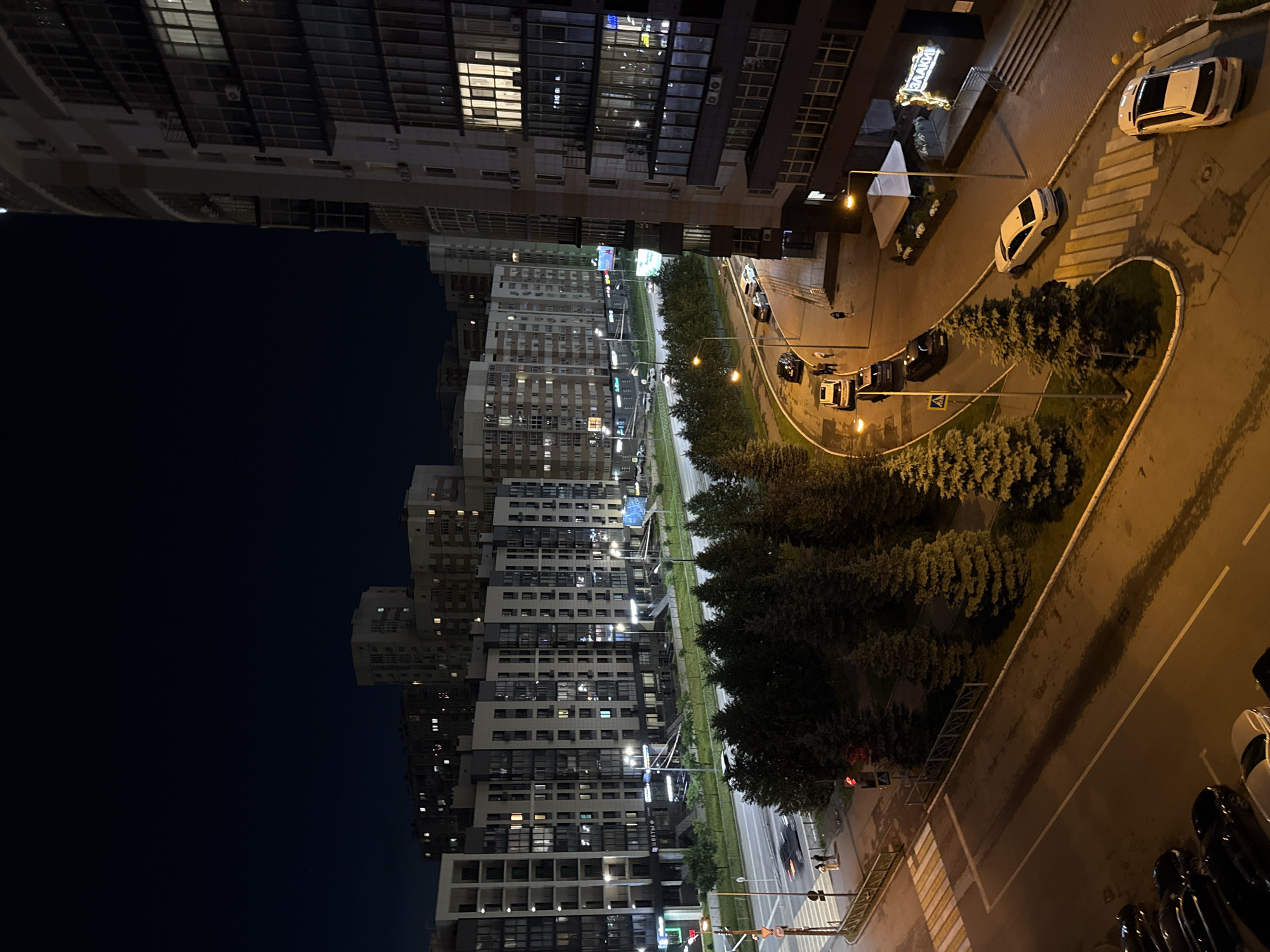 This is a night aerial shot of a street in Kazan, Russia, featuring a modern residential complex. The image captures a street scene with several cars parked and driving. On the left side of the image, a multi-lane road runs alongside a row of tall, contemporary apartment buildings with many illuminated windows. Adjacent to the road, a line of dense green coniferous trees forms a natural barrier. In the foreground, a crosswalk is visible, with two pedestrians standing on the sidewalk next to a traffic light. On the right side of the image, a taller, more complex building with a curved facade and multiple balconies dominates the scene. Below this building, a street-level entrance with a lit sign is visible. The overall atmosphere is urban and nocturnal, illuminated by streetlights and building lights.