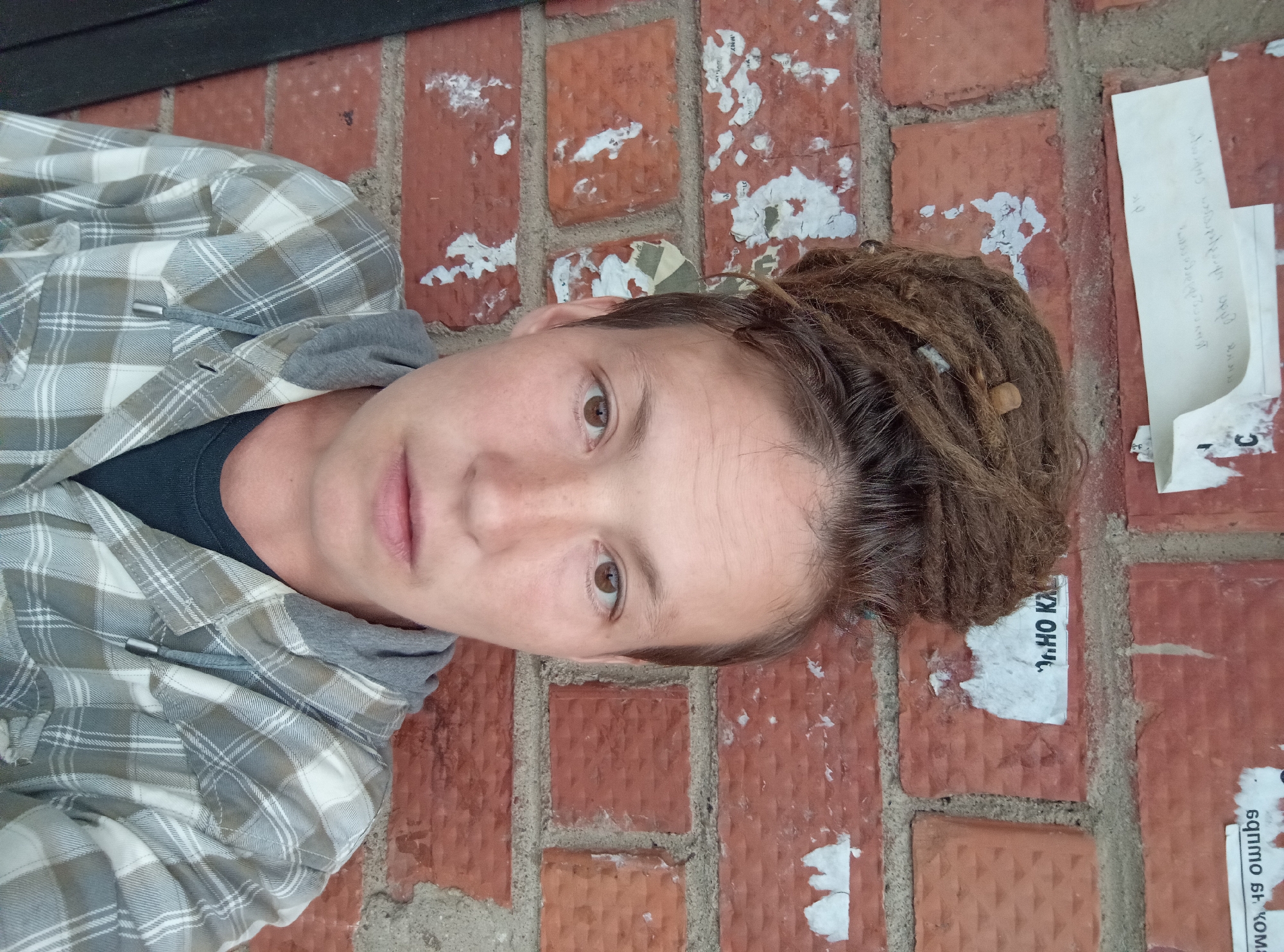 A close-up selfie shows a person with dreadlocks piled on top of their head, standing against a brick wall with torn posters. The person is wearing a gray and white plaid shirt over a dark shirt. The background consists of red bricks with various pieces of torn paper adhered to them. The lighting is natural and appears to be from the front, casting soft shadows. The overall scene suggests an urban outdoor setting.