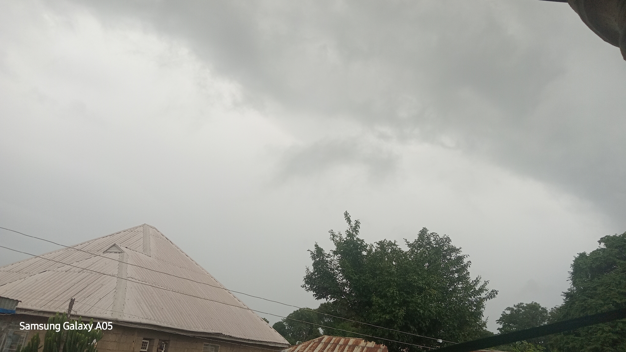 The image depicts an overcast sky above a residential area in Ribina East, Nigeria. The foreground features the sloped, corrugated metal roof of a building, its pale surface reflecting the diffused light. A triangular vent is visible near the peak of this roof. To the right, a dense canopy of green trees partially obscures the view, with some branches reaching into the frame. Another corrugated metal roof, this one showing signs of rust, is partially visible in the lower right corner. Power lines traverse the scene horizontally. The sky is filled with heavy, grey clouds, suggesting an impending storm or recent rainfall. The overall impression is one of a subdued, possibly humid, environment. No people are visible in the image.