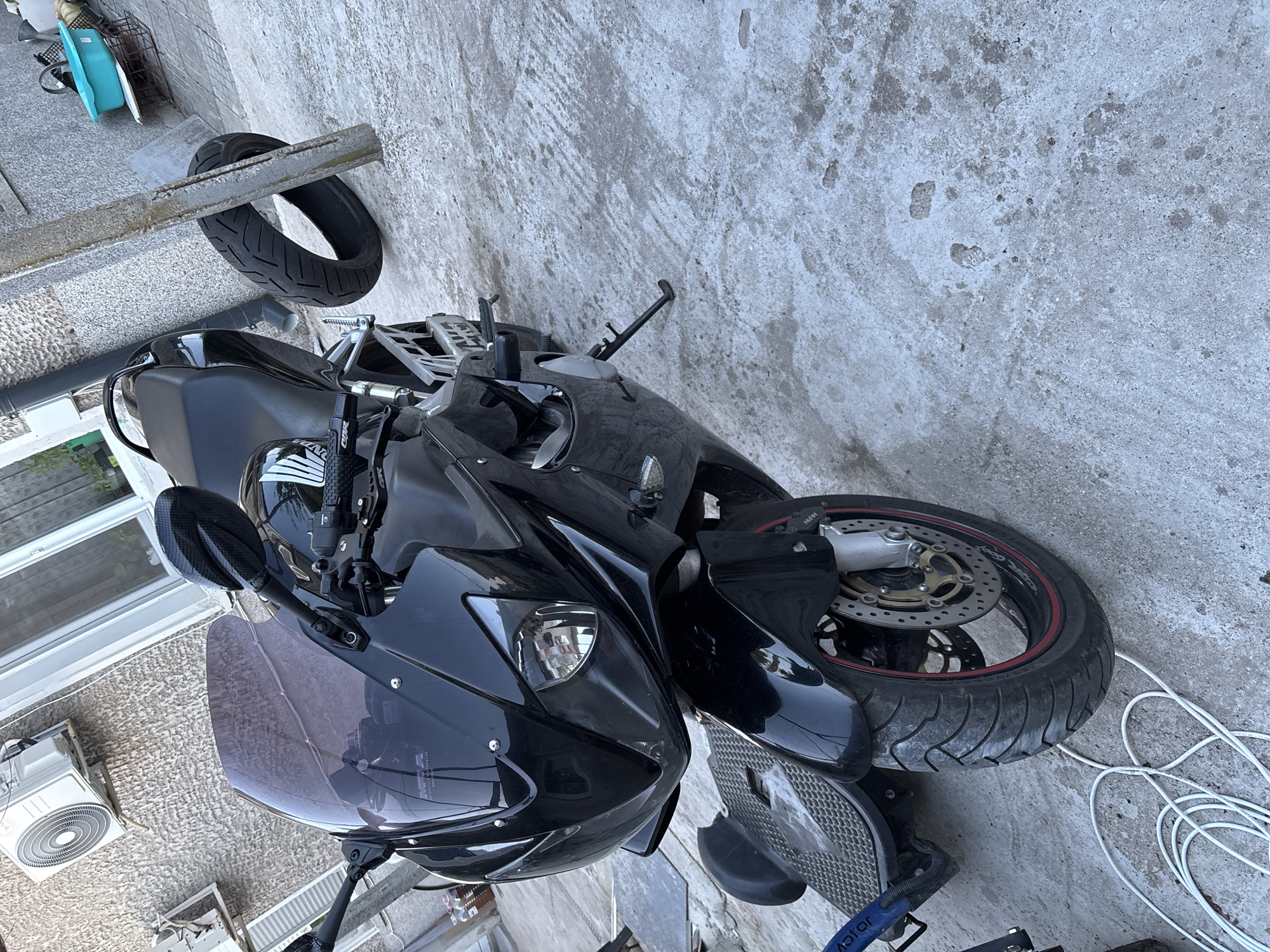 The image shows a black motorcycle parked on a concrete surface. A spare tire rests on a metal bar above the motorcycle. In the background, there's a building with an air conditioning unit. White wires are scattered on the ground near the motorcycle's front tire. The location is Unknown, Unknown.