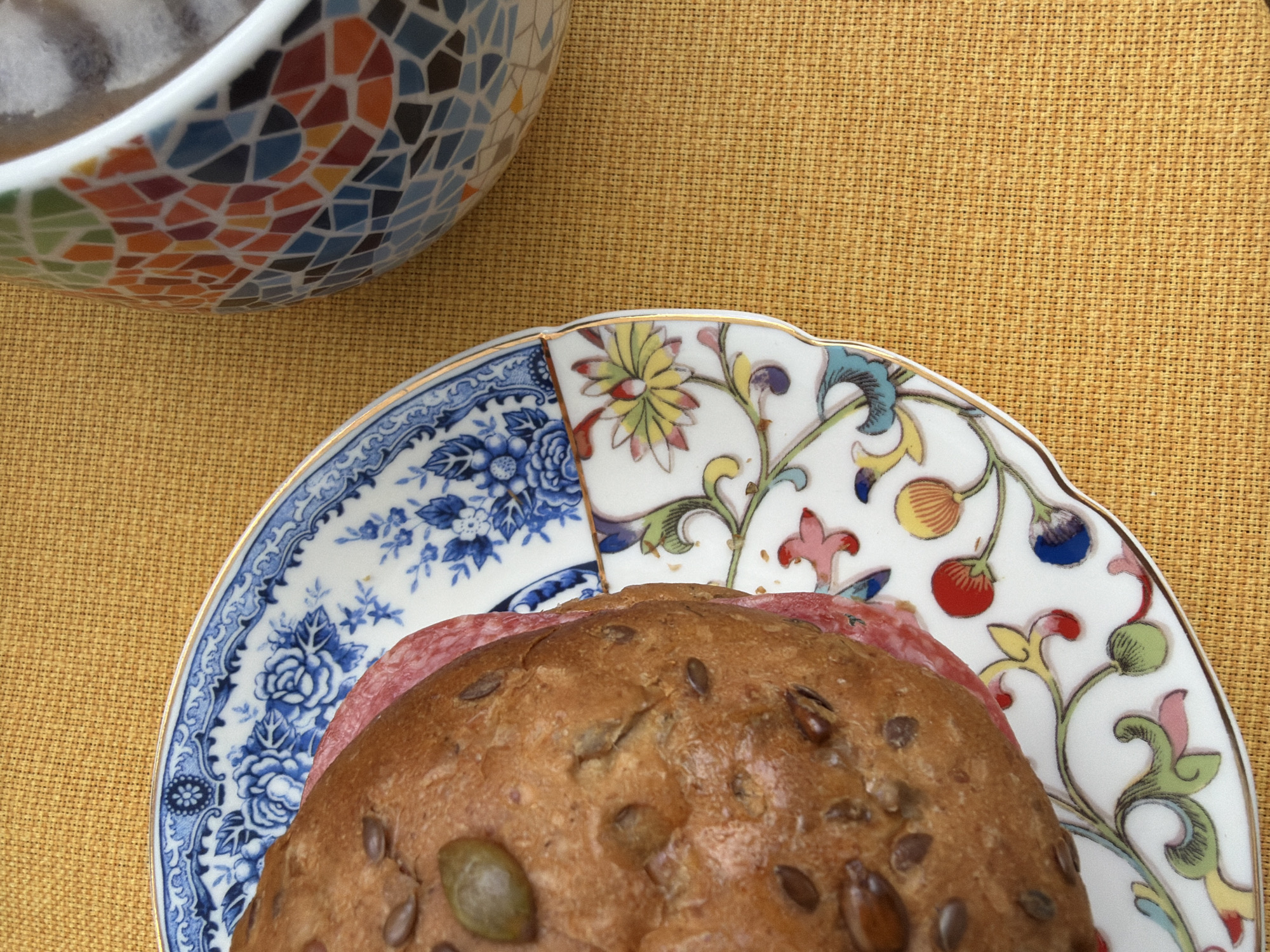 The image shows a sandwich on a decorative plate, next to a mosaic-patterned bowl, set on a textured yellow surface. The sandwich consists of a brown, seedy roll with a slice of salami inside. The plate has two different decorative patterns, one blue and white floral and the other floral with colorful accents. The mosaic-patterned bowl is in the upper left corner of the image. The location is not discernible from the provided visual information.