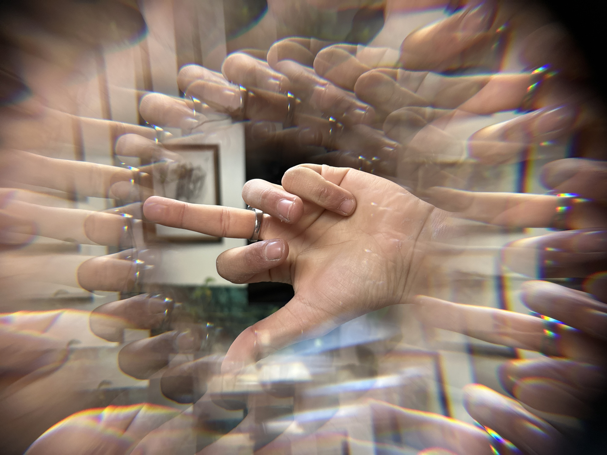 A hand making a "V" sign, wearing a silver ring, is captured in the center of the frame. The image is distorted with a multi-prism effect, replicating the hand and fingers multiple times in a radial pattern extending outward from the center. A framed picture is visible in the background. Based on metadata, the photograph was taken in Palaiseau, France.