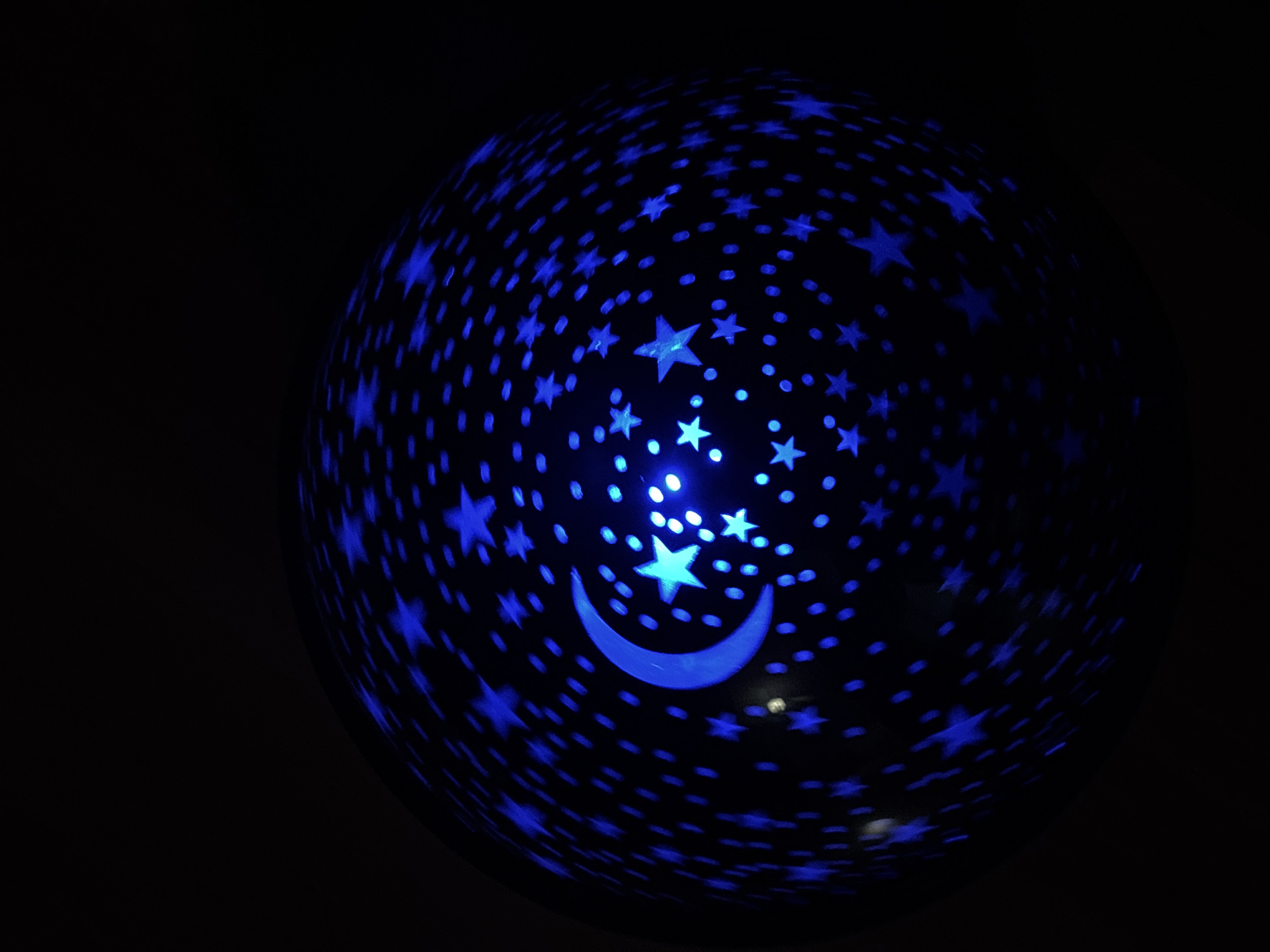A close-up shot shows a dark sphere with blue star and crescent moon patterns on its surface. The sphere appears to be emitting light, giving the patterns a glowing effect. The background is entirely black, emphasizing the illuminated sphere. There is no indication of a location in Borino, Bulgaria, or any other information about people, subjects, objects, actions, or interactions beyond the described sphere and background.