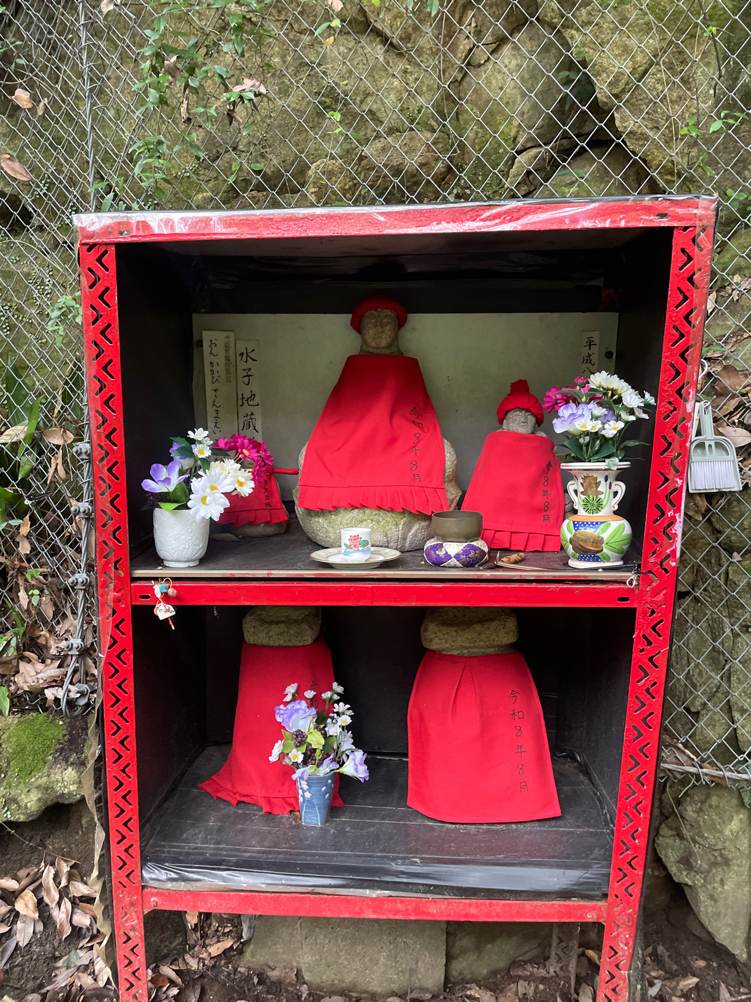 This is a medium shot of a red, open-fronted, multi-shelf shrine structure with a wire mesh fence and a rocky, wooded hillside in the background. The shrine contains several stone statues of what appear to be Jizo Bodhisattvas, figures from Japanese Buddhism. The statues are dressed in red cloth garments, and some wear red hats. There are offerings of flowers in vases and what appear to be small bowls. A small dustpan and brush are hanging on the right side of the shrine. The shrine is located outdoors, with fallen leaves and mossy rocks in the foreground. The overall scene suggests a spiritual or devotional site, possibly a roadside shrine or a place of worship in a natural setting. The image lacks any discernible details that would pinpoint a specific location within Kobe, Japan.