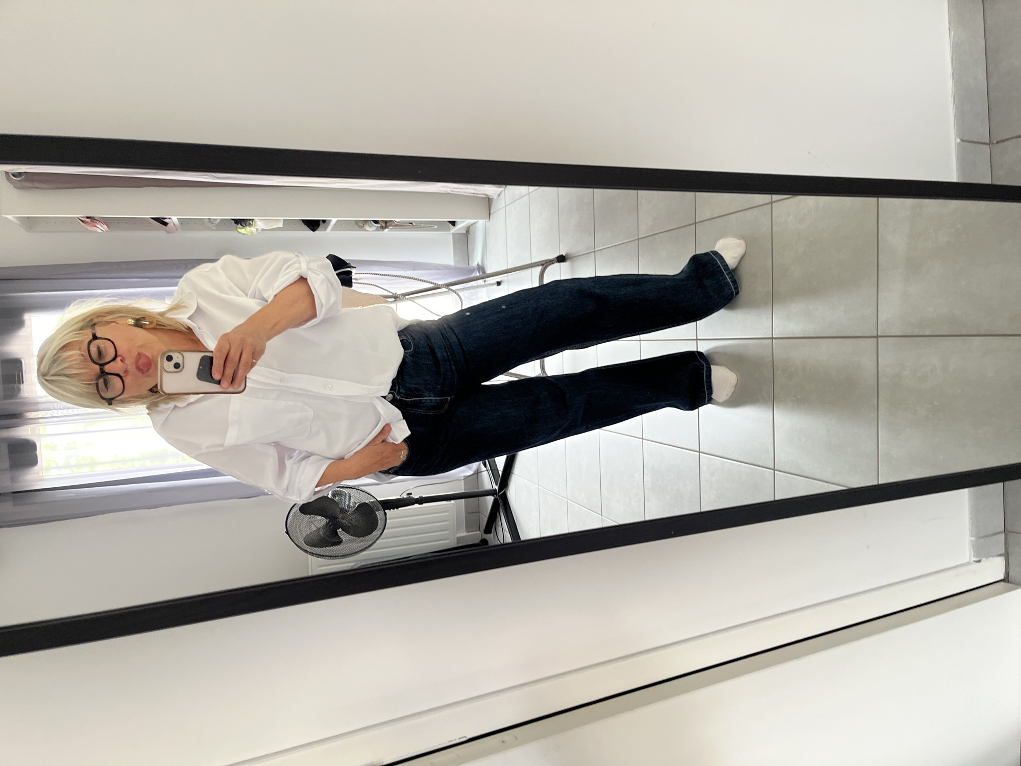 The image is a mirror selfie taken by a fair-skinned woman with blonde hair, wearing glasses, a white collared shirt, and dark denim jeans. She is standing indoors, likely in a bedroom or dressing area, based on the presence of a wardrobe or shelving unit in the reflection. A portable fan is visible in the background, along with a chair or a small foldable ladder. The floor is tiled with light gray square tiles. The reflection suggests the room is well-lit, possibly by natural light coming through a window with a curtain. The location may be Chambœuf, France, but this cannot be confirmed from the image alone.