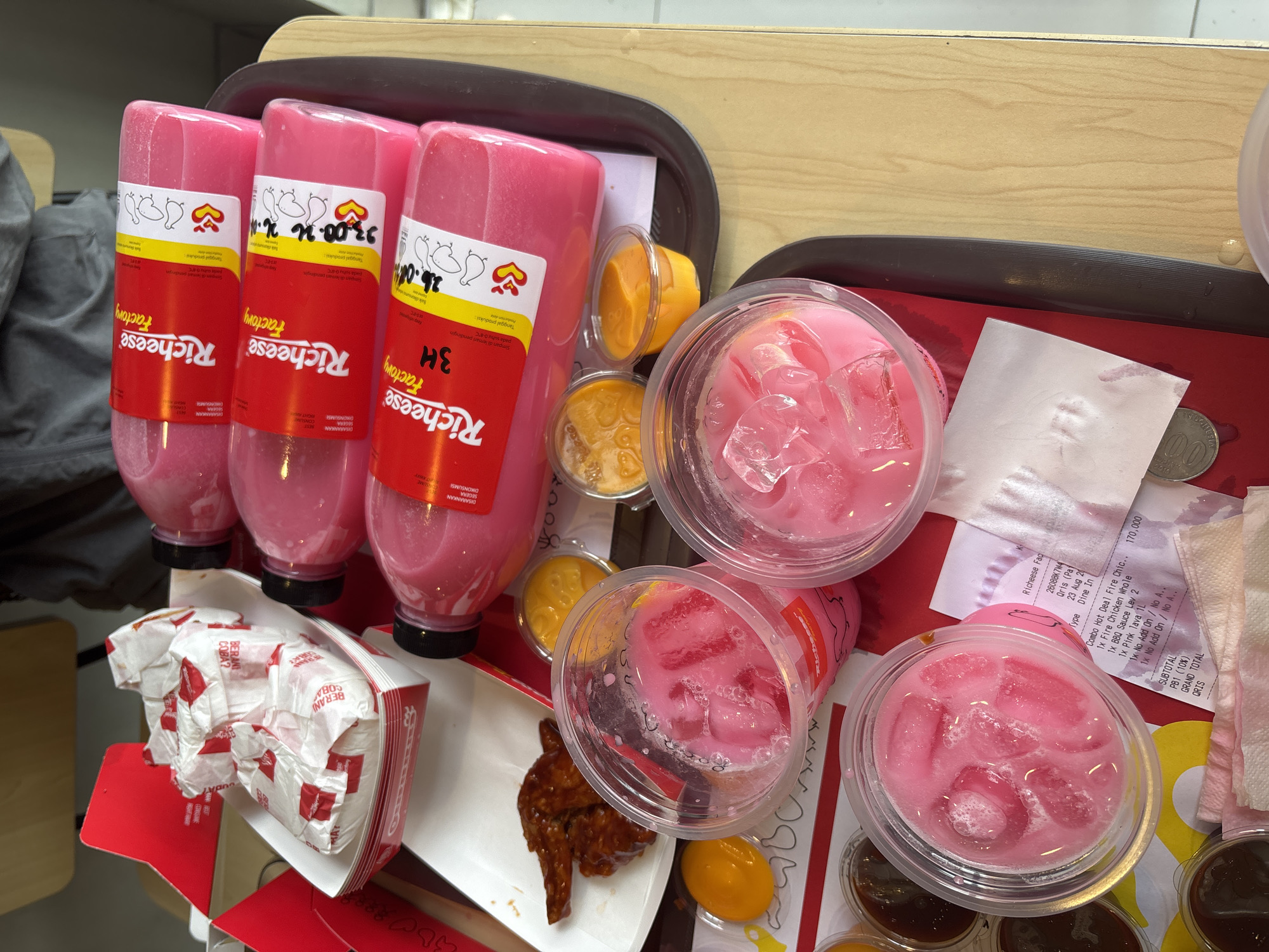 A close-up overhead shot shows a tray of Richeese Factory food items and drinks on a light brown wooden table. The tray, which is dark grey with a ribbed edge, holds several food containers and drinks. In the center-left, a white paper tray contains several pieces of fried chicken wings coated in a dark brown sauce, wrapped in white paper with red branding. To the right of the chicken, four pink plastic bottles are stacked in a row. Each bottle has a red label with white text that reads "Richeese Factory" and features cartoon ducks. Three of the bottles have additional yellow labels with dates and numbers. In front of the bottles, there are three small clear plastic cups containing a bright yellow sauce, and another two clear plastic cups filled with a pink beverage and ice. A crumpled receipt is visible between the pink drinks and one of the yellow sauce cups. The background shows a red and white Richeese Factory cardboard box and a portion of a person's grey jacket. The scene suggests a dine-in or takeout order from a Richeese Factory restaurant.
