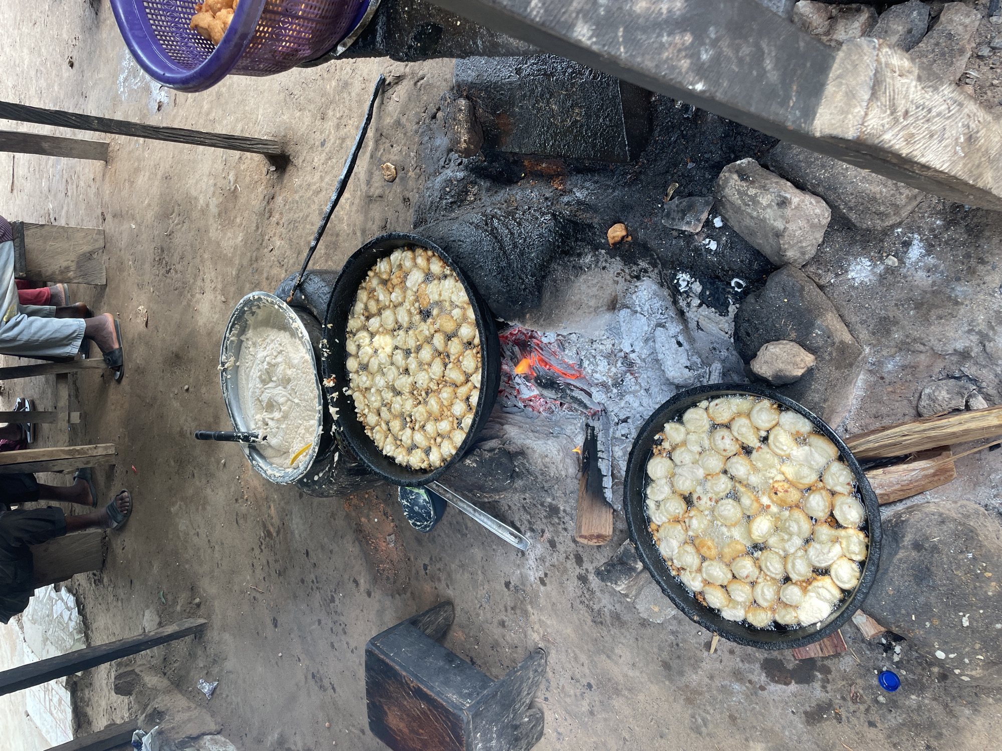 The image depicts an outdoor cooking setup, likely in Keffi, Nigeria. Two large, black pans containing numerous small, round dough balls are frying in oil over an open fire, fueled by wood and charcoal. A third metal pot holds a white batter-like substance, possibly the mixture used to make the dough balls. Utensils are visible near the pots, suggesting active cooking. The cooking area is surrounded by rocks and ash.
A person is partially visible, seated on a wooden structure, with only their lower legs and feet in sandals shown. A purple basket filled with cooked dough balls is in the upper left corner. The ground is a mix of dirt and concrete.