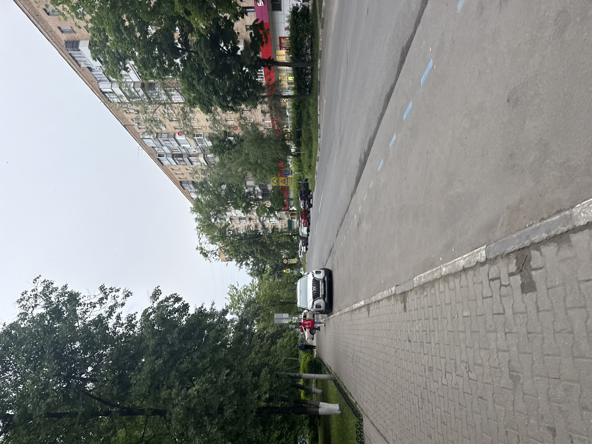 The image shows a street scene in Mytishchi, Russia. The camera is positioned on the sidewalk, looking along a road lined with trees and a tall residential building. The building, typical of Soviet-era construction, features multiple balconies and rows of windows. A white car is parked along the sidewalk, and a person wearing a red shirt and dark pants is walking near the car. Several other people are walking further down the sidewalk. The road is gray asphalt with blue markings. Street signs are visible in the distance. The sky is overcast. The sidewalk is paved with gray rectangular paving stones. A metal fence borders the sidewalk.