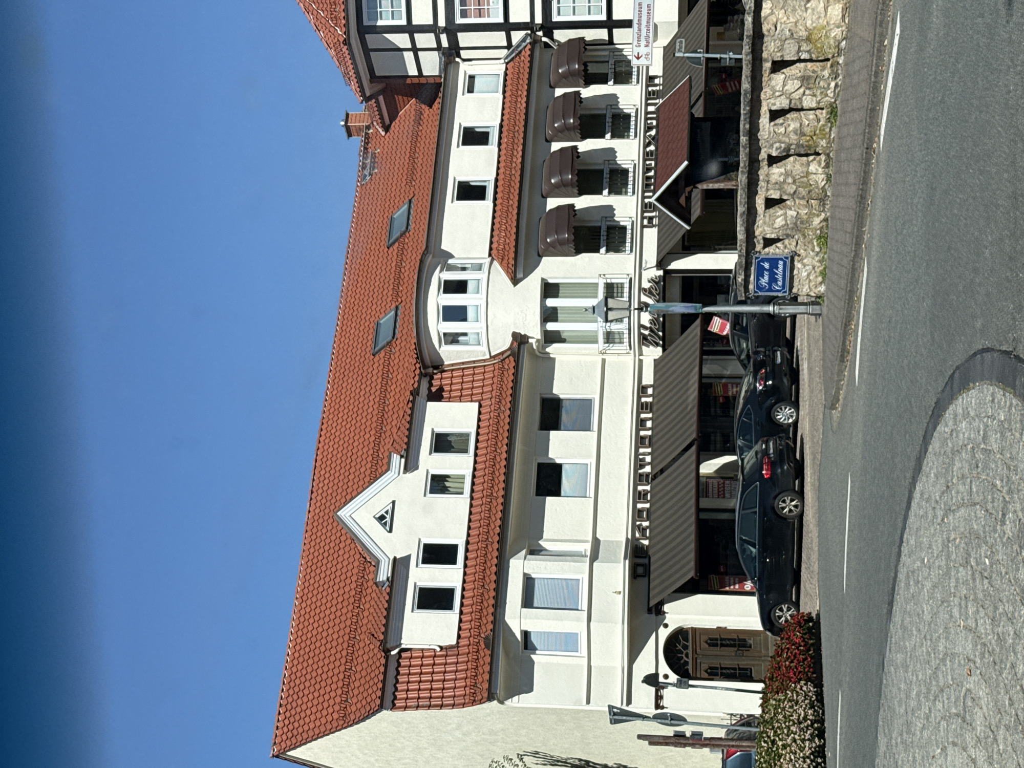 A white, multi-story building with a red tile roof and several windows dominates the frame. The building appears to be a commercial establishment with awnings extending over the ground-floor windows and doors. The signage above the awnings is partially obscured but includes the words "DER KORSETTHAUS" and "WEILER". To the right of the main building, another building with half-timbered detailing is visible. A stone wall with triangular cutouts runs along the sidewalk in front of the building. A street sign indicates "Grenzlandmuseum" and "Naturzeitmuseum," suggesting the location is in a town with historical and natural attractions. Two black cars are parked in front of the building, and a street lamp stands on the sidewalk. The scene is set against a clear blue sky. The area appears to be a town square or a commercial street in Bad Sachsa, Germany.