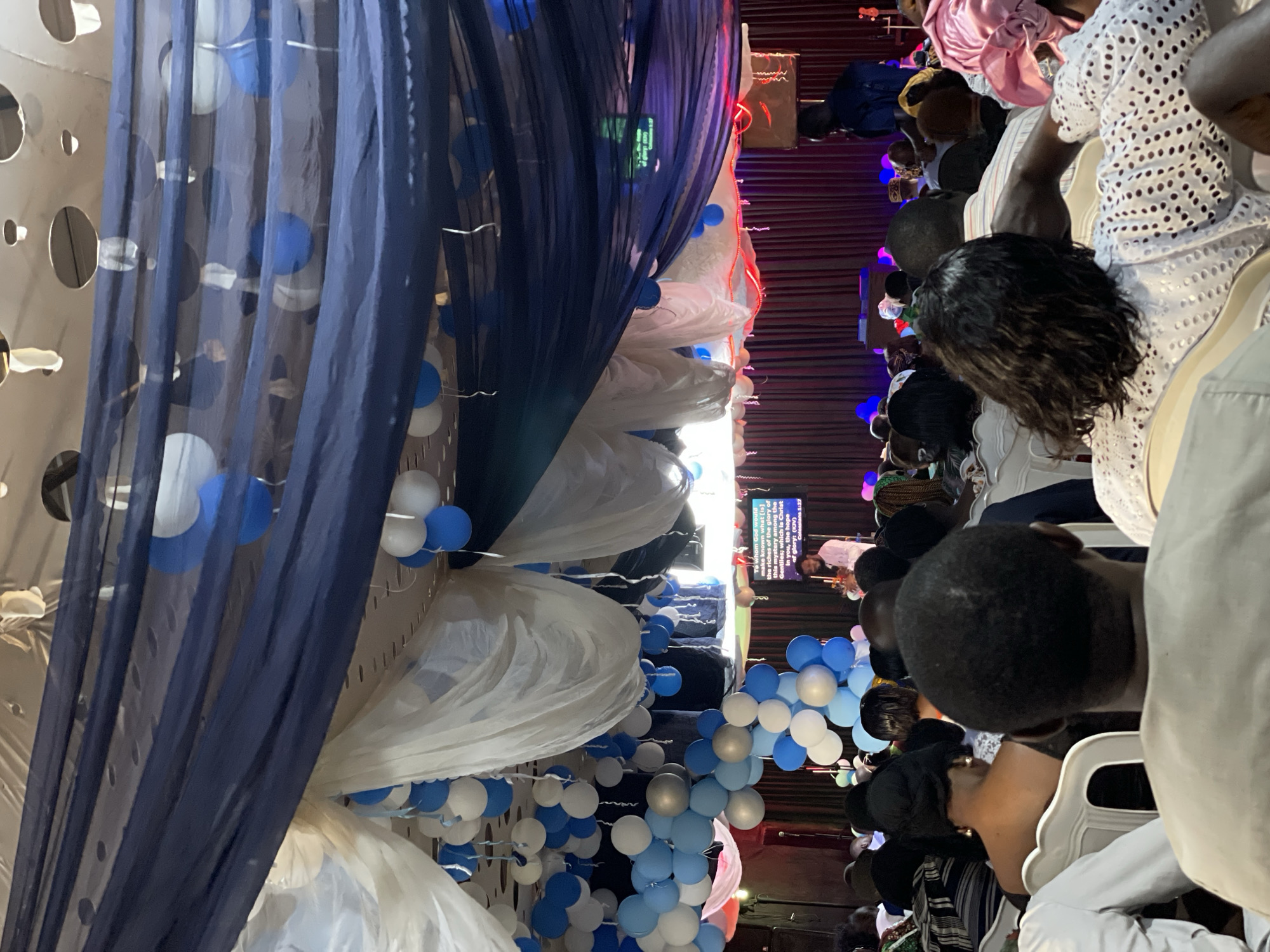 The image depicts an indoor gathering, likely a church service, held in Durumi, Nigeria. The scene is decorated with numerous blue and white balloons suspended from the ceiling, along with draped fabrics in dark blue and white. A small screen displays text, possibly a scripture verse. Several individuals are visible, predominantly in the foreground, seated and facing a stage area. Their attire suggests a formal or semi-formal occasion. In the background, a person stands on a stage, seemingly addressing the congregation. The overall atmosphere appears to be one of community and worship.