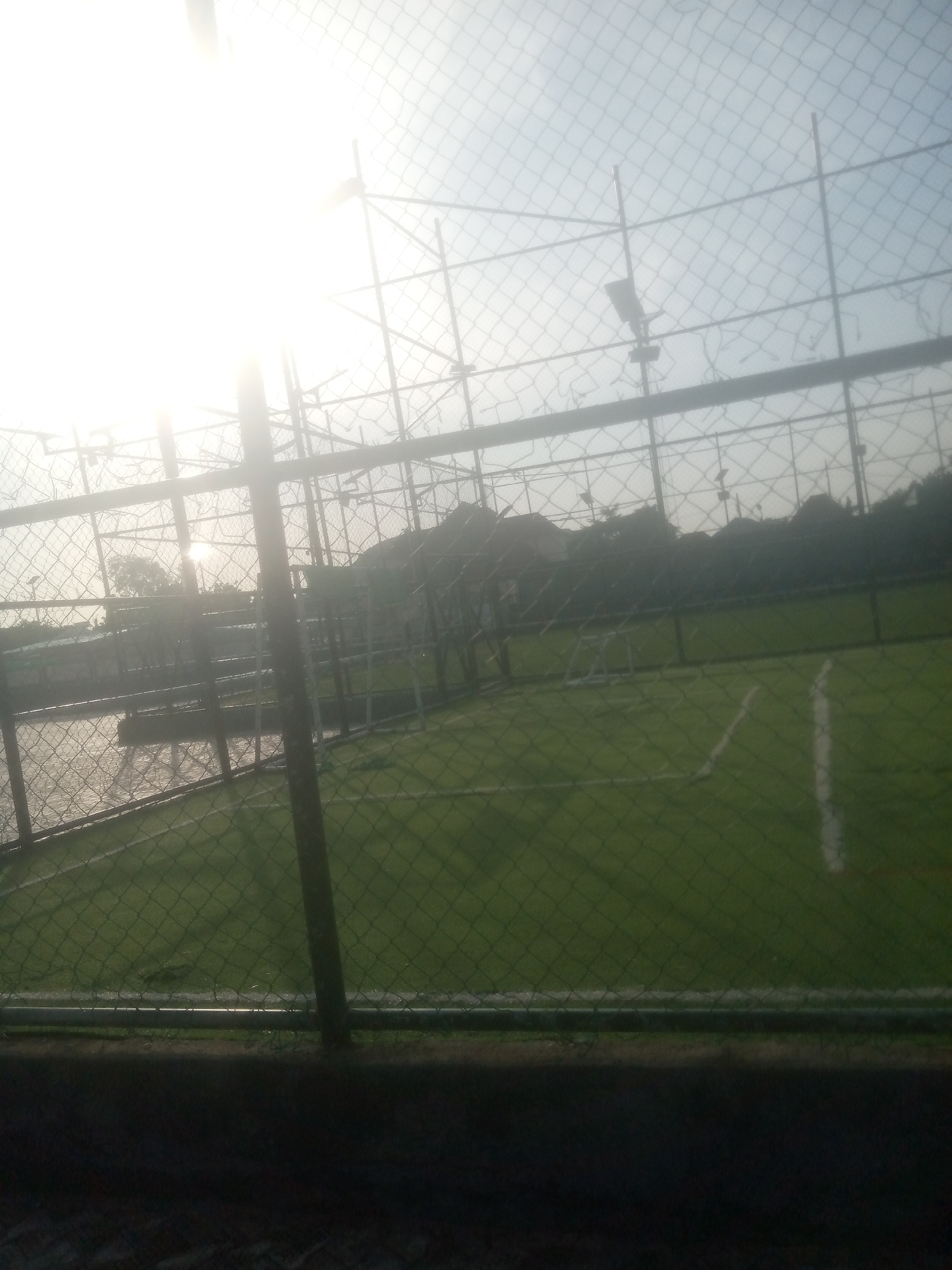 The image depicts an outdoor sports facility, likely a futsal or small-sided soccer field, enclosed by chain-link fencing. The field itself is a vibrant green, with white lines marking boundaries and a central area. The sun is low in the sky, creating a bright glare that washes out parts of the scene, particularly in the upper portion of the image. This suggests the photograph was taken during either sunrise or sunset, casting long shadows and a diffused light across the field.
The fencing is prominent throughout the frame, with a large, dark vertical pole in the foreground on the right side. Through the netting, glimpses of trees and what appear to be buildings can be seen in the background, indicating an urban or semi-urban environment. There are also metal structures, possibly part of the lighting system or additional fencing, visible above the field. The ground outside the fenced area appears to be a mix of concrete and possibly a darker, rougher surface in the immediate foreground.
No people are visible in the image, nor is there any clear indication of a specific event taking place. The overall mood is quiet and still, accentuated by the natural light and the empty sports venue. The location context of Jalingo, Nigeria, suggests a warm climate, which aligns with the appearance of an outdoor sports field. The weather appears clear, despite the intense sunlight. There is no visible text in the image.