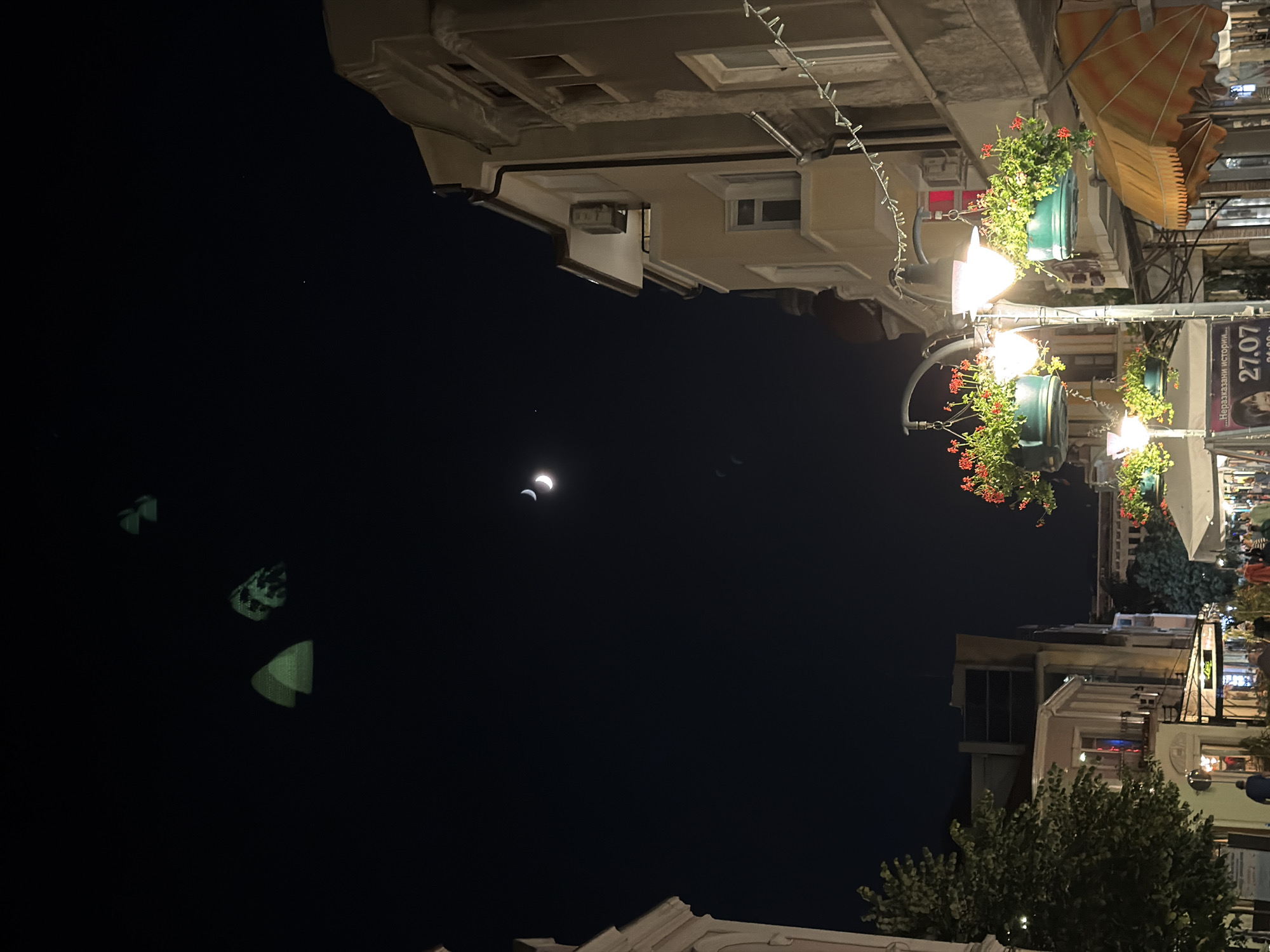 The image shows a street scene at night in Burgas, Bulgaria, likely on a pedestrian street. The sky is dark, with a crescent moon visible and a few stars. Three faint, green, triangular shapes are in the upper left portion of the sky, possibly reflections or lights from an event. The street is lined with buildings, some appearing to be shops or restaurants, with awnings and lights. Several street lamps are present, each adorned with hanging baskets of red flowers. People are walking along the street, though their faces are not clearly discernible. There are also some trees and foliage visible. In the background, there are more buildings and lights, suggesting a developed urban area. A poster with text, including a date "27.07", is visible on a stand. The overall impression is a lively evening atmosphere in a European city.