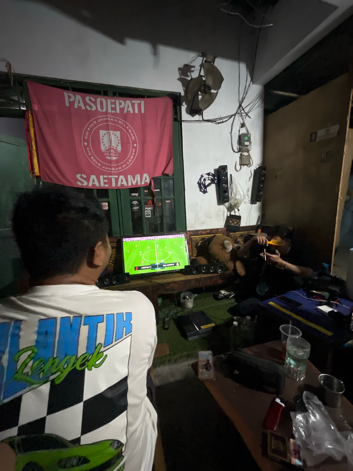 The image depicts an indoor scene, likely in a dimly lit room within Desa Gagaksipat, Indonesia. In the foreground, a person is seen from behind, wearing a white long-sleeved shirt with a checkered pattern and graphics of a green car and text that appears to say "LANTIK Zengek". This individual is facing a television screen displaying a soccer game.
The room contains several objects, including a red banner with text and a logo hanging on the wall above the television. The banner displays "PASOEPAIT" in large letters and a circular emblem with text and a depiction of a rocket and stars. Below this, "SAETAMA" is also visible.
To the right of the television, a teddy bear is positioned on a surface. Further to the right, another person is seated, holding what appears to be a comb and a cigarette, and looking towards the left. Various electronic devices, including speakers and a power strip, are present in the room.
On a table in the foreground, there are several items such as a mobile phone, a dark-colored bag, disposable cups with liquid, and what looks like a metal cup. The overall environment suggests a casual gathering, possibly for watching a sports event. The lighting is low, with shadows contributing to the atmosphere.