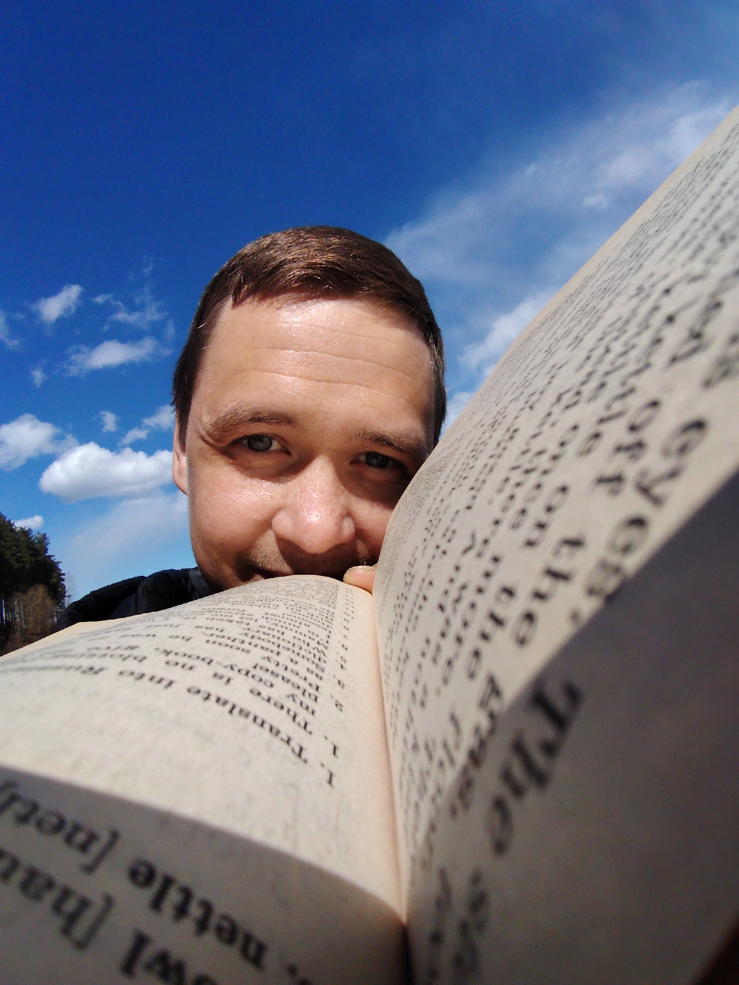 A close-up, low-angle selfie captures a man peering over an open book outdoors in Chaikovsky, Russia. The man, with short brown hair, fair skin, and blue eyes, looks directly at the camera with a subtle, friendly smile. He holds the open book in front of his face, partially obscuring the lower half, suggesting he is reading or posing while taking the picture.
The book's pages are filled with printed text, primarily in English, with some words and phrases clearly visible despite being slightly out of focus. Identifiable text includes "eyes," "off the," "nettle," and a section starting with "I. Translate into Ru" (Russian). This section lists numbered phrases like "1. There in," "my copy-book," "please!", "Pretty soon," "Dante," and "Somebody," indicating a language exercise or learning material.
The background features a vivid, clear blue sky dotted with scattered white cumulus clouds, indicating fair weather during the daytime. On the far left, a distant line of dark green trees suggests a natural, possibly forest, setting. The lighting is bright and natural, highlighting the details of the man's face and the book's pages.