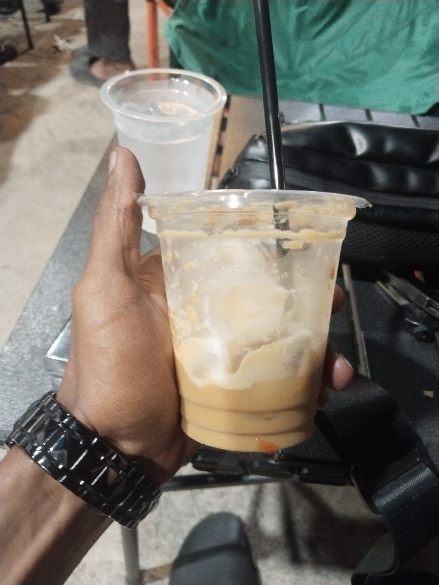 A person holds a partially consumed iced coffee in a clear plastic cup with a black straw, near another clear plastic cup filled with clear liquid. The person wears a black watch on their left wrist. A black bag with a strap is visible in the background, along with various objects including what appears to be scissors. The setting seems to be a casual outdoor seating area, possibly in the city of Lhokseumawe, Indonesia. The lighting is moderate. Other people are partially visible in the background.