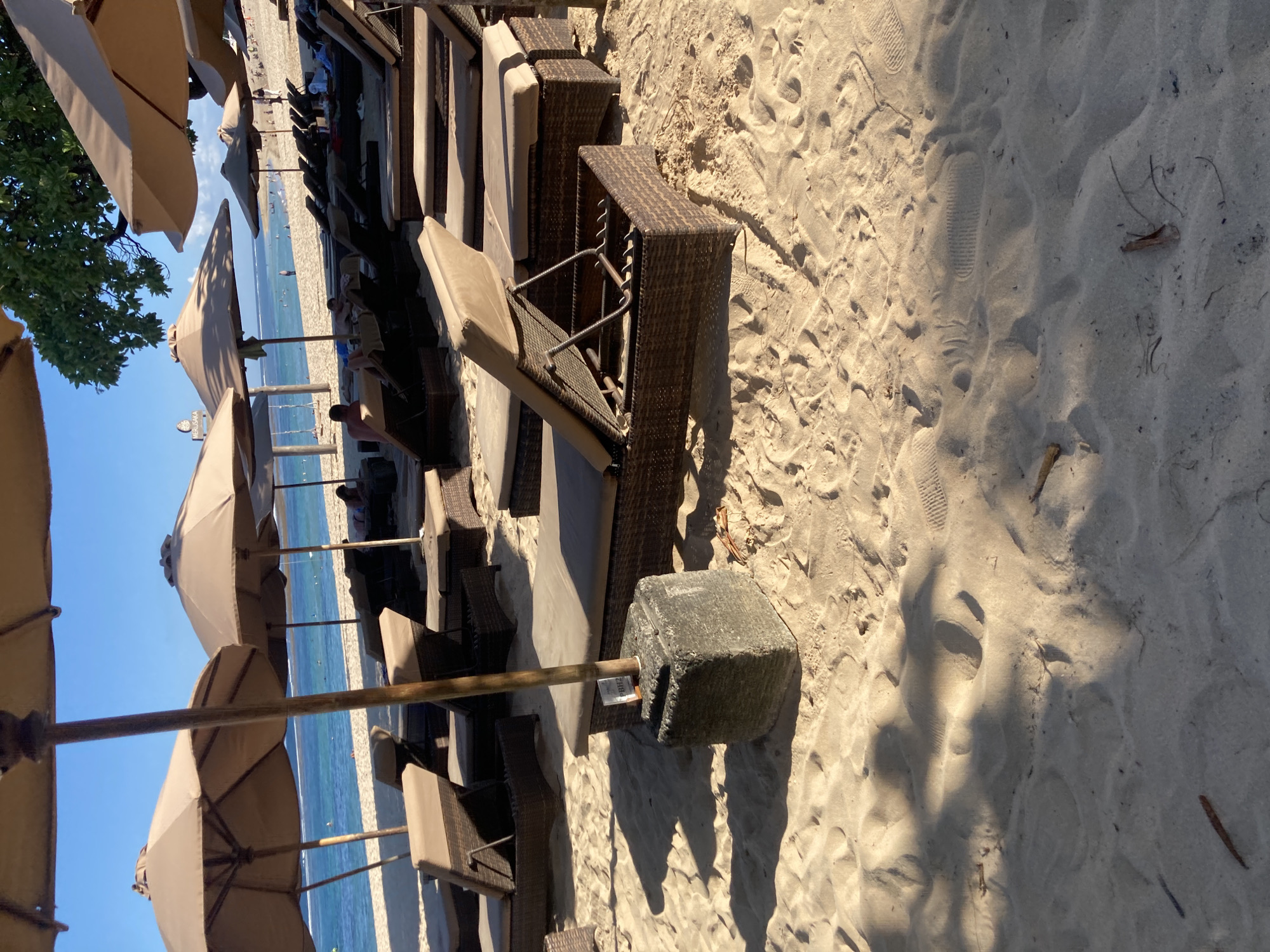 The image depicts a beach scene in an unknown location. Rows of beach chairs and umbrellas line the sandy shore, with the ocean visible in the background. The sand is light-colored and shows footprints. The umbrellas are tan and block the sun. The beach chairs are made of wicker and have beige cushions. A large, concrete block anchors one of the umbrellas. The scene appears to be a resort or private beach, with no visible people.