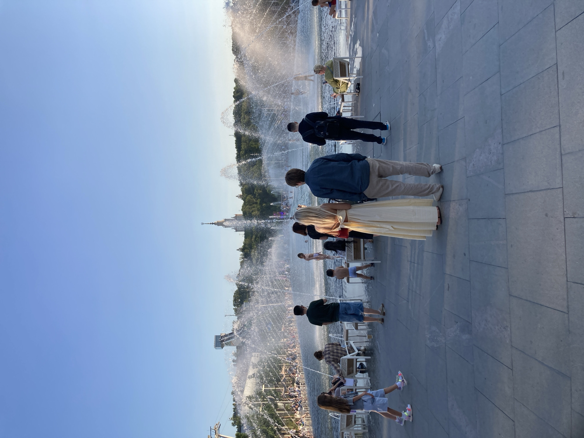 The image depicts a wide fountain with numerous water jets spraying into the air. In the foreground, a couple stands with their backs to the camera, facing the fountain. The woman wears a long, flowing cream-colored dress, and the man is in a blue button-down shirt and beige pants. Other people are scattered throughout the scene, some sitting at tables and chairs near the fountain, while others stand or walk. In the background, a large building with a spire, resembling the Moscow State University skyscraper, is visible against a clear blue sky. Trees line the horizon behind the fountain. This scene is likely located at the VDNKh (Exhibition of Achievements of National Economy) park in Moscow, Russia, known for its grand fountains and architecture.
