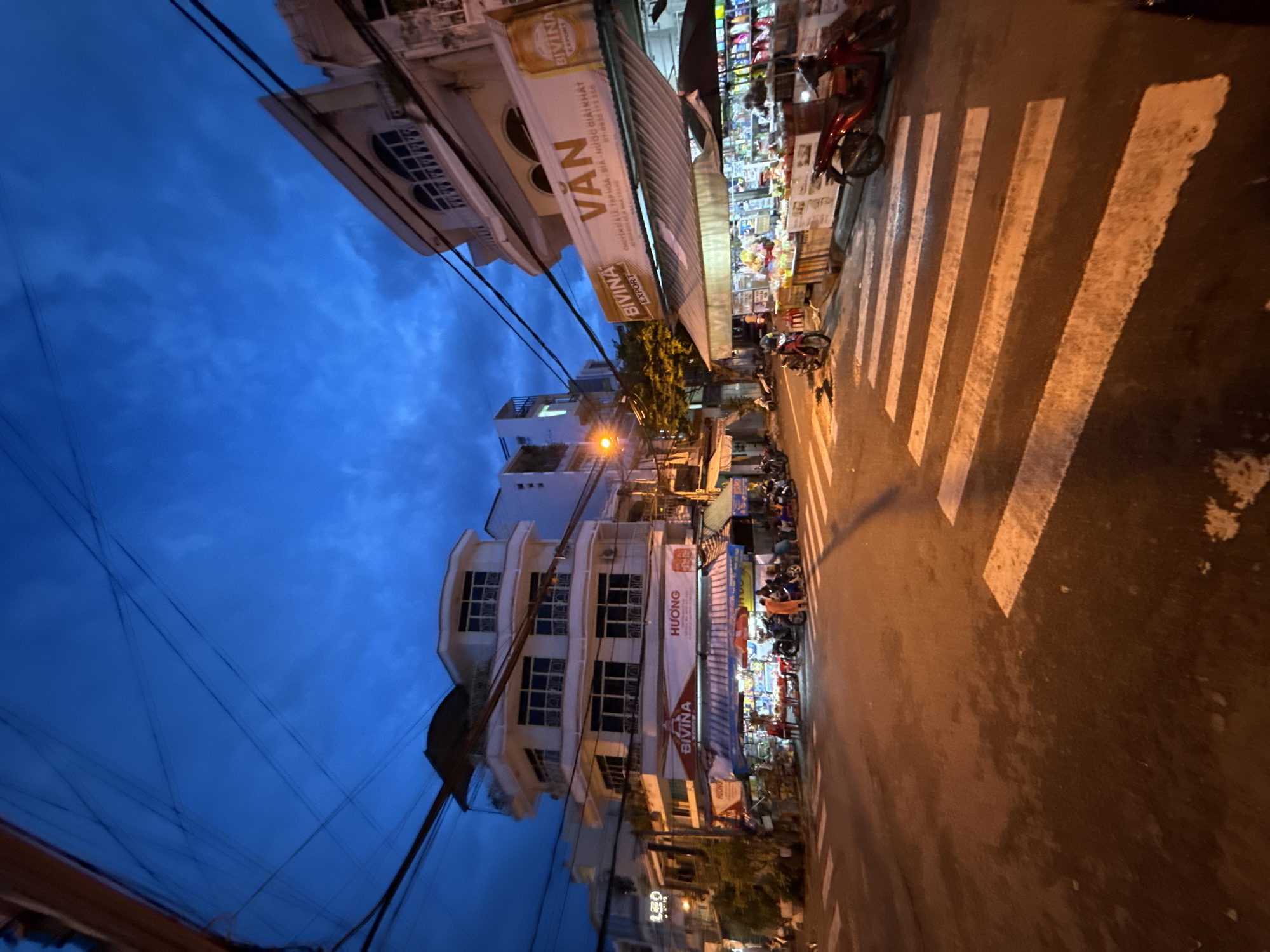 This is an exterior, ground-level, wide-angle shot of a street corner in Nha Trang, Vietnam, taken at dusk. The sky is a deep, vibrant blue with scattered clouds. A complex network of overhead wires crisscrosses the upper portion of the frame.
In the foreground, a pedestrian crossing is clearly visible on the asphalt road. To the right of the crossing, two red scooters are parked. Beyond the crossing, shops line the street. One shop has a large awning displaying the brand name "BIVINA" in red and white letters, and another awning advertises "VĂN" in large white letters on a brown background, with "BIVINA EXPORT" also visible. The shops appear to be selling various goods, with shelves and displays visible behind the awnings.
Across the street, a multi-story white building with multiple balconies and windows dominates the left side of the image. Several other buildings of varying heights and architectural styles are visible behind it. People are present on the sidewalks and in front of the shops, some appearing to be customers or vendors. A few individuals are interacting, and some are standing or sitting. The overall atmosphere suggests a busy commercial area in the early evening.