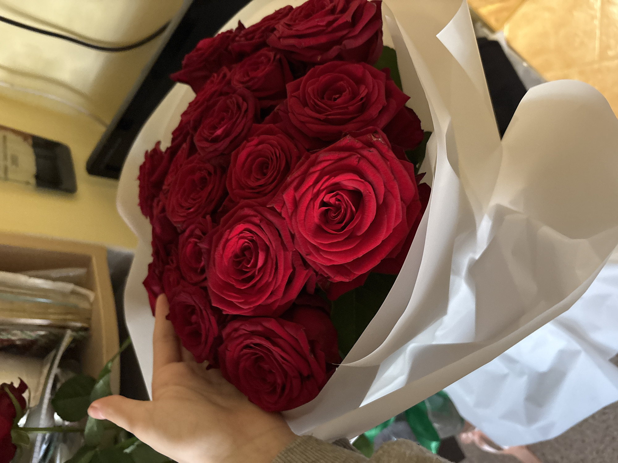 A person's hand, with light-toned skin and a green sweater sleeve, is holding a bouquet of red roses wrapped in white paper. The roses are in full bloom and tightly packed. The background is blurred, showing a yellow wall and some shelves with items on them. The scene appears to be indoors. There is no information about the location in Minsk, Belarus.