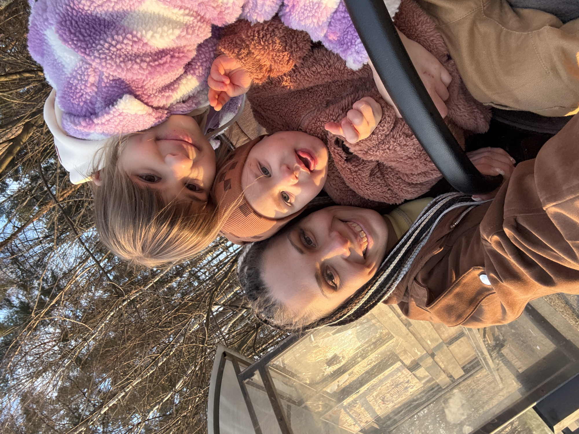 A woman and two young children are close together, looking up at the camera. The location is outdoors with trees in the background. The image is oriented so that the ground and trees are above the subjects. The woman is at the bottom of the frame, smiling and wearing a brown jacket. She has braided hair. To the left of the woman is a young girl with blonde bangs and a purple fleece jacket. A baby in a brown fleece outfit is in the center. The location is not recognizable as Rusanovo, Russia.