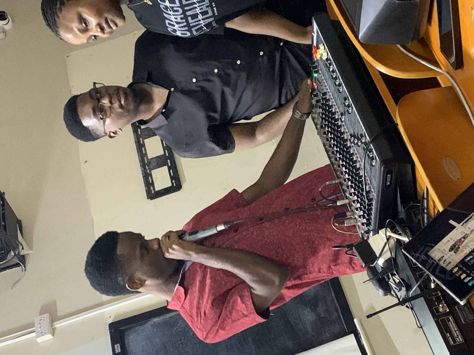 The image depicts an indoor setting where three individuals are engaged in what appears to be a recording or audio production session. A man wearing a red collared shirt is holding a microphone and speaking into it. Beside him, another man is operating a mixing console, wearing glasses and a black button-down shirt. A third individual is partially visible, wearing a black t-shirt. Various pieces of audio equipment are present, including speakers, cables, and a tablet, set up on what looks like a wooden desk or workstation. The environment suggests a recording studio or a space adapted for audio production. There is no information to ascertain if the photo was taken in Dar es Salaam, Tanzania.