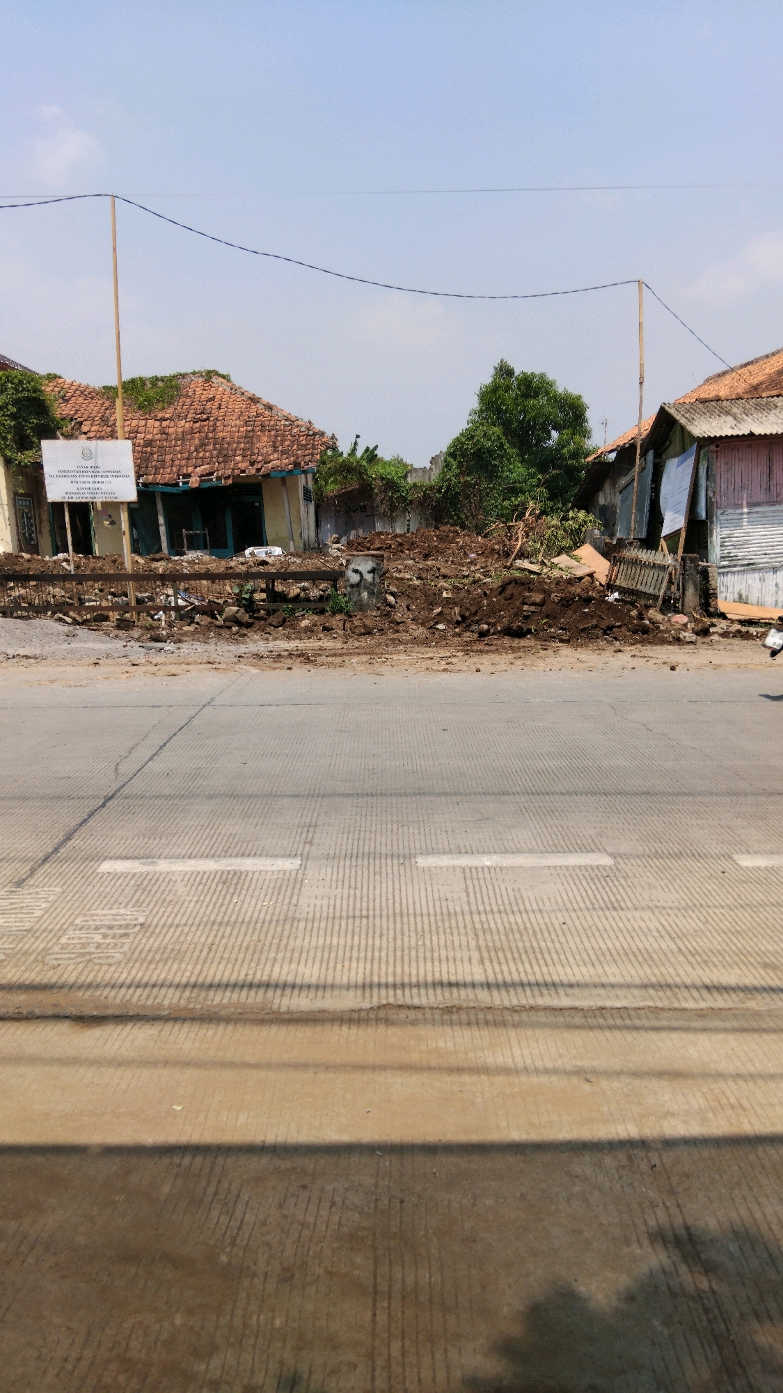 The image shows a scene of demolition or construction in the city of Batang, Indonesia. In the foreground, a paved road with a textured surface is visible, with some debris and shadows on it. In the background, there are partially demolished or damaged buildings with orange tiled roofs. A large pile of soil and rubble sits in front of these structures, suggesting ongoing work. A white sign with text in Indonesian is prominently displayed to the left of the debris. Telephone wires are strung across the scene, and trees are visible behind the buildings. The sky is clear and blue. The overall impression is one of a site undergoing significant change or repair.