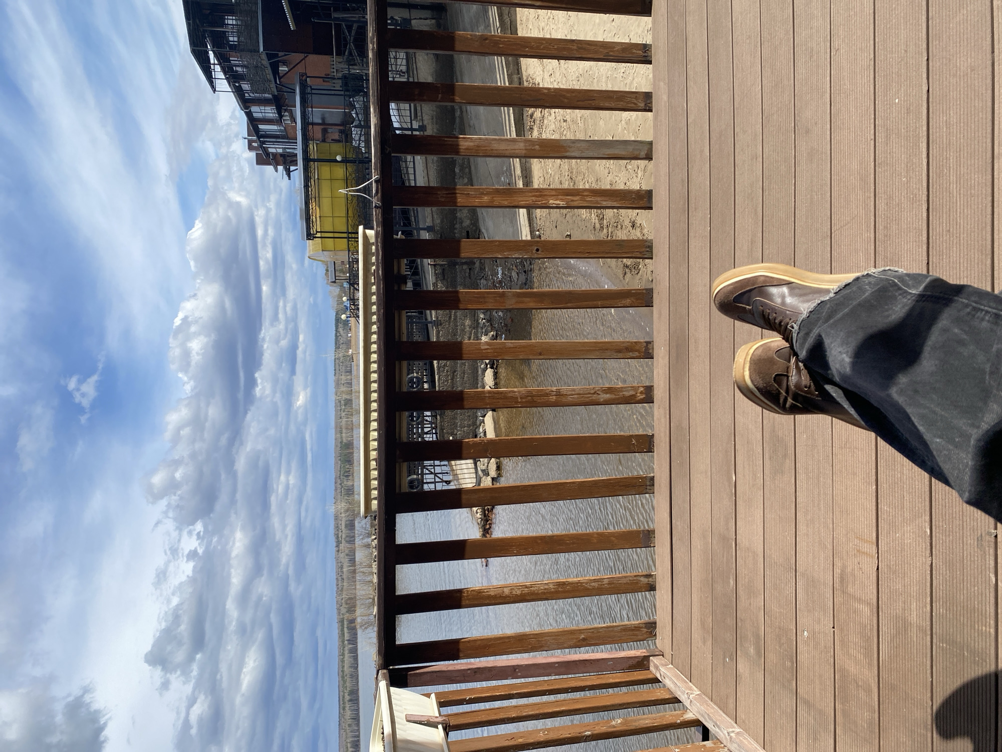 The image depicts a first-person perspective from a wooden deck or pier, overlooking a body of water and distant buildings under a partly cloudy sky, likely in Chaikovsky, Russia.
In the foreground, the lower legs and feet of a person are visible, wearing dark blue/black jeans and dark brown boots with light tan soles. The person is resting on a light brown wooden deck, which features parallel grooves. Strong sunlight casts a distinct shadow of the person's legs and feet onto the deck.
A wooden railing, constructed from horizontal and vertical dark brown slats, separates the deck from the view beyond. Through the railing, a calm body of water is visible, reflecting the bright sky. On the opposite bank, several multi-story buildings can be seen, some with modern architecture and one featuring a bright yellow awning. The distant shoreline beyond the buildings appears to have sparse trees or vegetation.
The sky is a bright blue, filled with numerous white and grey, streaky clouds, indicating a partly cloudy day. The scene is brightly illuminated by natural daylight, suggesting it is either morning or afternoon, given the clear and strong shadows. There is no visible text in the image. The overall impression is one of peaceful observation of the surrounding landscape from a relaxed vantage point.