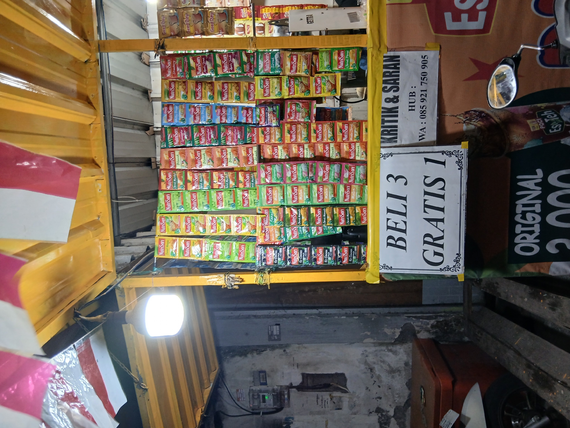 The image shows a street stall in Denpasar, Indonesia, at night. The stall is illuminated by a bright lightbulb hanging from the yellow awning. Shelves display a large assortment of colorful packets of NutriSari, a popular Indonesian powdered drink mix, in various flavors. To the right of the NutriSari display, there are packets of "Good Day" coffee. A sign on the stall reads "BELI 3 GRATIS 1" (Buy 3 Get 1 Free) and another sign provides a WhatsApp contact number for "KRITIK & SARAN" (Complaints & Suggestions). In the background, a concrete wall with peeling paint and an electrical meter are visible. A motorcycle and a cooler are also present in the foreground. The overall scene depicts a typical roadside vendor operating after dark.