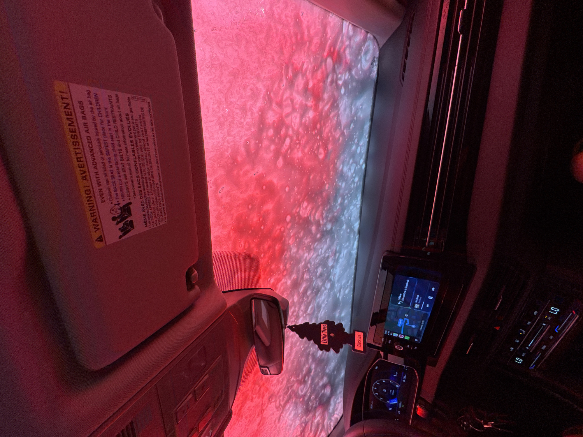 The image depicts the interior of a vehicle, observed from a low angle, while actively undergoing an automated car wash in Roselle, United States of America. The exterior view through the windshield and sunroof is dominated by thick, red-tinted foamy water and numerous white bubbles, indicative of a washing cycle. The red light emanating from the car wash facility heavily illuminates the car's interior.
Inside the vehicle, the dashboard is visible, featuring a digital instrument cluster on the left displaying numerical values and a blue graphic accent. A central infotainment screen shows a navigation interface with a map, a turn instruction in Spanish ("Da vuelta a la izquierda" - Turn left), and music playback information ("Un Beso"). Below the screen, the climate control panel displays "LO" on both driver and passenger sides. A black "Little Trees" air freshener, labeled "Black Ice," hangs from the rearview mirror, which is centrally positioned. The passenger-side sun visor is flipped up, revealing a yellow and white warning sticker with text in English and French regarding "ADVANCED AIR BAGS" and "CHILD RESTRAINTS," emphasizing the back seat as the safest for children. A portion of the steering wheel is visible on the left, and a set of keys hangs near the lower left dashboard area. No individuals are visible within the frame.