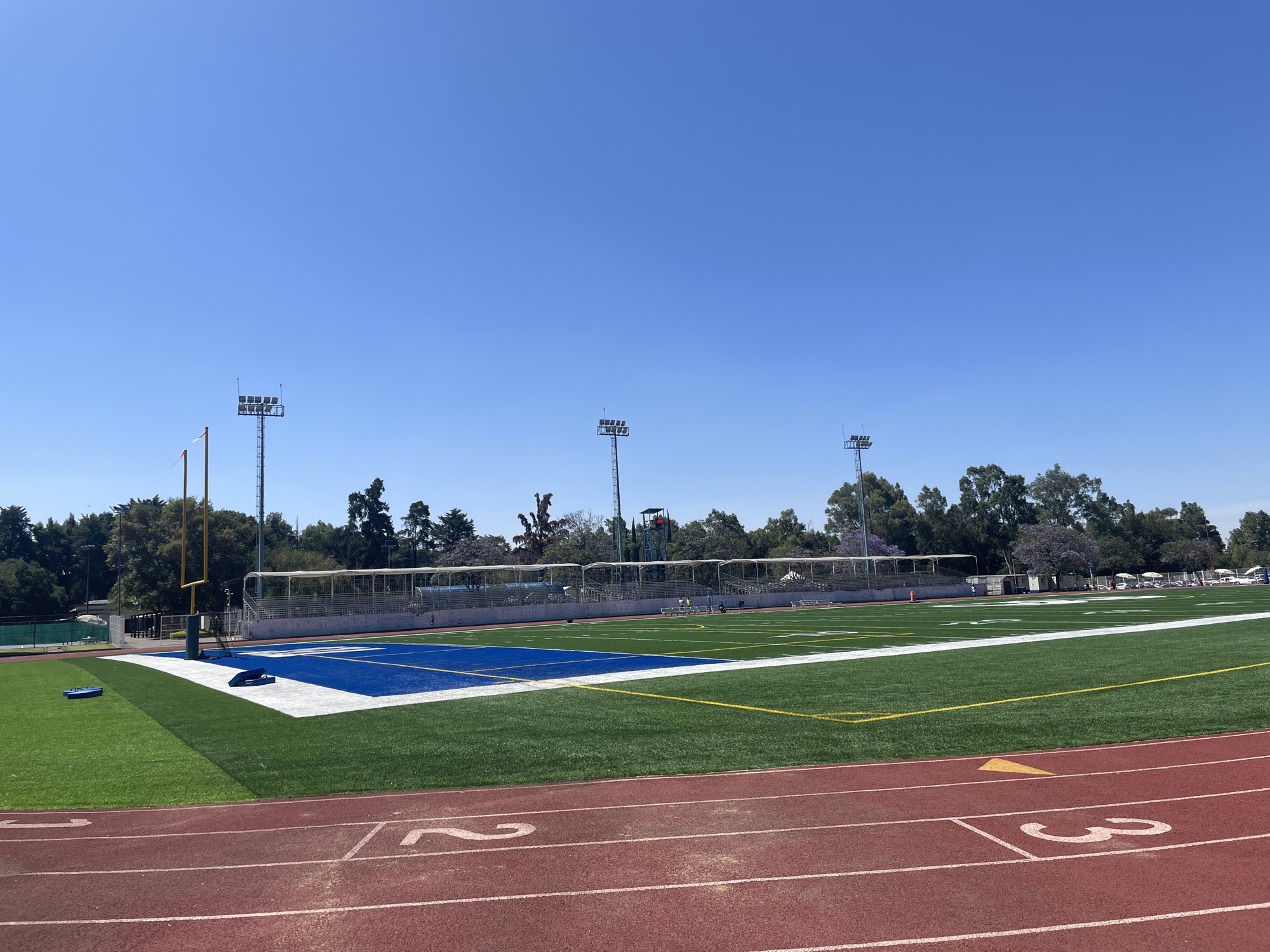 The image presents a view of a sports field with an athletic track in the foreground. The track has a red surface and numbered lanes. The field itself is green, with white and yellow yard lines indicating its use for football or a similar sport. One endzone is colored blue. In the background, there is a covered grandstand or bleacher area, behind which trees and foliage are visible. Tall light posts are stationed around the perimeter of the field. The sky is clear and blue.
Given the landscape, architecture, and general appearance, the image is likely from the "Deportivo Chapultepec" located in Mexico City, Mexico.