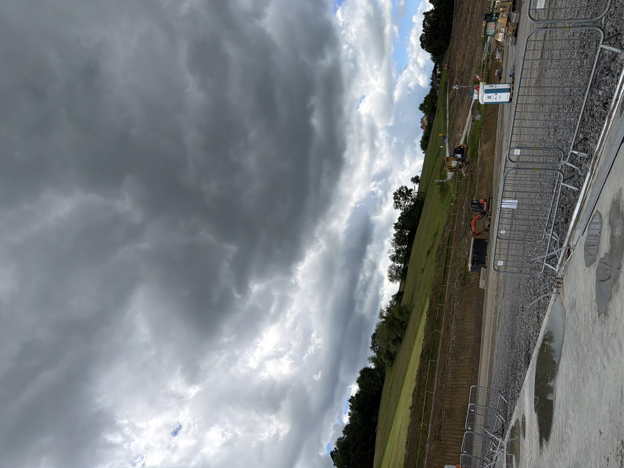 The image depicts a construction site in a rural area with a hill in the background. The sky is filled with dramatic, dark storm clouds. In the foreground, there are several metal safety barriers and puddles of water on a concrete surface, indicating recent rain. The construction site itself is characterized by piles of gravel, a portable toilet, and heavy machinery, including an excavator and a bulldozer. A large black container is also visible. The background features a grassy hillside dotted with trees and a few buildings, suggesting a residential area or scattered houses. The overall impression is one of a vast open space undergoing significant development under an impending storm.