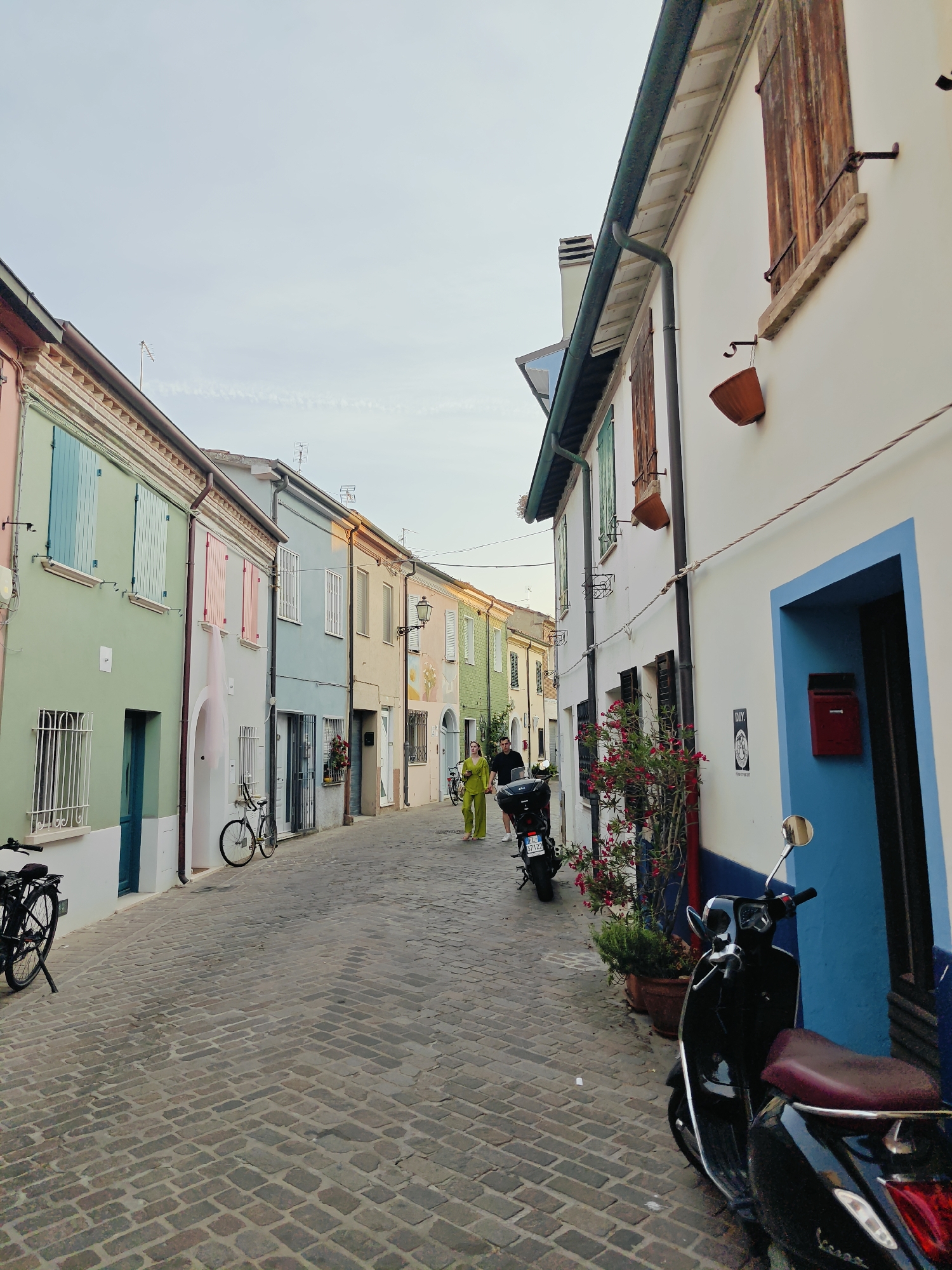 The image depicts a charming, narrow cobblestone street lined with brightly colored houses in Rimini, Italy. The buildings feature pastel facades in shades of pink, green, blue, yellow, and cream, with contrasting shutters and doors. On the right side of the street, a white building with a vibrant blue doorway and a red mailbox is prominent. A black Vespa scooter with a burgundy seat is parked in front of this building.
In the middle ground of the street, a woman wearing a bright yellow jumpsuit walks away from the camera. Another person, partially obscured, is walking behind her. A bicycle is parked on the left side of the street. The street is paved with grey cobblestones, and potted plants with red flowers add a touch of color.
The scene appears to be set in the late afternoon or early evening, indicated by the soft, warm light casting subtle shadows and the overall gentle illumination. The sky is a pale blue with wispy clouds, suggesting fair weather. The architecture, including the shutters, window designs, and the style of the houses, is characteristic of Italian coastal towns. The presence of bicycles and scooters suggests a pedestrian-friendly and leisurely environment. The overall mood is peaceful and serene, evoking a sense of a quiet, picturesque Italian town.
A license plate is visible on the back of the Vespa, reading "P 57122". A small black and white sign with the text "D.I.Y." is affixed to the white wall on the right.