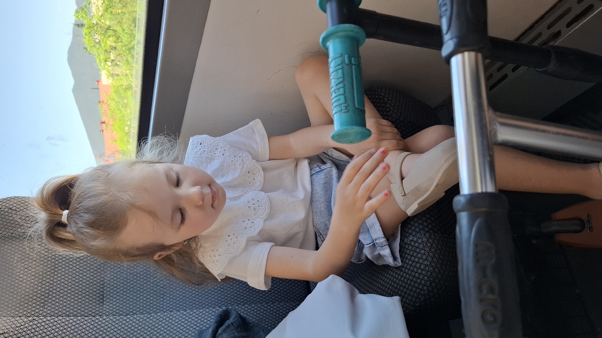 A young child is resting in an upright seated position within the interior of what appears to be a vehicle. The child has light skin and light hair pulled into a ponytail with a white band. She wears a white shirt with a lace collar, denim shorts, and light tan shoes. Her fingernails are painted a shade of pink.
The setting is inside a vehicle, possibly a bus or train, as indicated by the gray seat and a view out the window showing greenery and buildings, possibly in a mountainous area. The child is positioned next to the window. Above and near her, there is a blue handle and a silver and black metal frame, part of an object such as a scooter, lying across the seating area. The location of this scene is Unknown, Unknown.