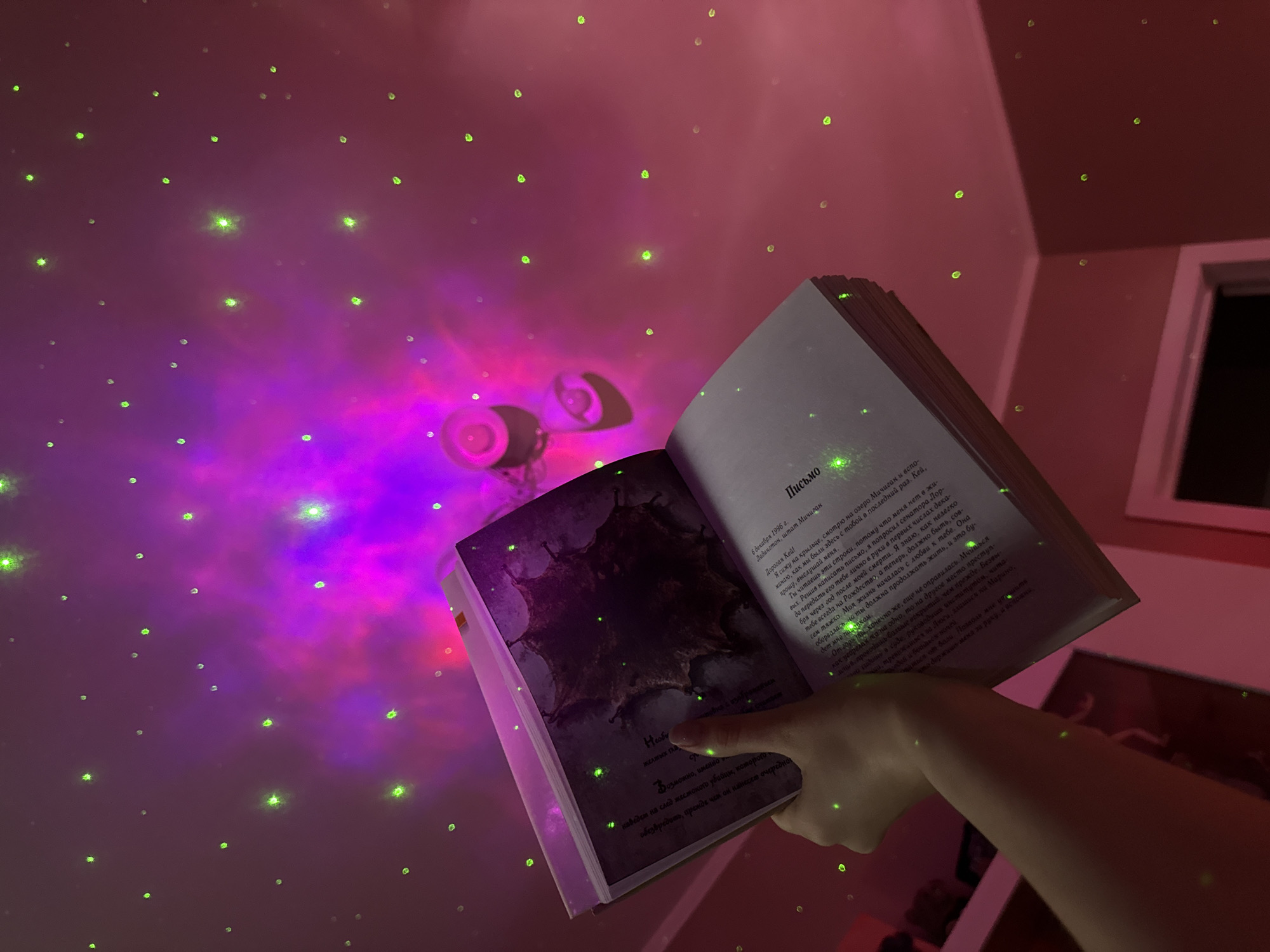 Here's a forensic description of the image:
**Location:** Appears to be an indoor setting, likely a residential room.
**Environment:** The room is illuminated by colored lighting, predominantly pink and purple, with green "star" projections on the walls and ceiling.
**Subjects:** A hand is holding open a book.
**Objects:**
* An open book, displaying text in Russian (likely the Cyrillic alphabet) and an image that appears somewhat grotesque or fantastical. The text includes the word "Письмо" (Letter).
* Wall-mounted light fixture on the ceiling with two adjustable lights.
* A window with what appears to be dark outside, and a shelving unit that is out of focus.
**Actions:**
* The hand is holding the book open.
**Interactions:**
* The book is being illuminated by the room's lighting.
**Details:**
* The green "star" projections are visible on the book pages as well as the walls.
* The text in the book seems to be the focus of the image.
* The light fixture projects colorful light onto the walls and ceiling.