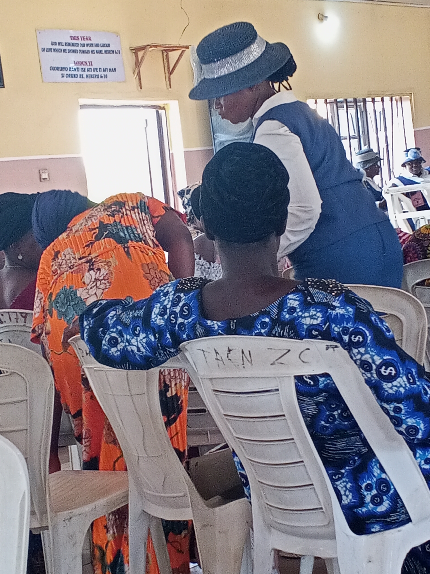 The image depicts the interior of a building, possibly a church or meeting hall, in Zango-ndaji, Nigeria. The foreground features several rows of white plastic chairs. A woman in a blue and white patterned dress sits in one, her back to the camera. The back of her chair is marked with "TAEN ZC." An adjacent woman is partially visible in an orange floral dress. Another woman, possibly an attendant, stands, wearing a blue suit vest over a white shirt and a blue and silver hat. Several other people are visible in the background, some also wearing hats. A sign hangs on the wall, with text in both English and another language, likely Yoruba, referencing God's remembrance of work and love. There is a window.