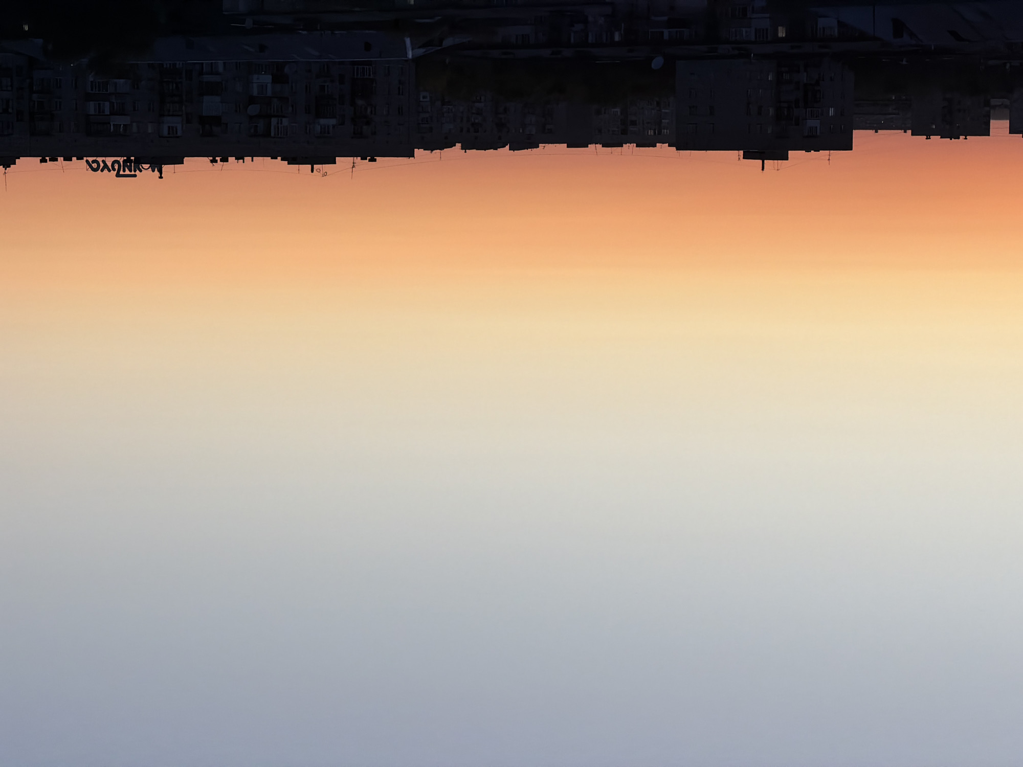 The image captures a panoramic view of apartment buildings in Pavlodar, Kazakhstan, during a sunset. The sky transitions from a soft blue at the top to a vibrant orange and yellow hue near the horizon, creating a gradient effect. Silhouetted against this colorful sky are rows of multi-story apartment buildings, their dark forms starkly defined. A prominent feature on the rooftop of one of the buildings is graffiti that appears to spell out "NOVAYA," a word meaning "new" in Russian. The overall scene is one of an urban landscape bathed in the warm glow of twilight, with the graffiti adding a touch of modern urban art to the architecture.