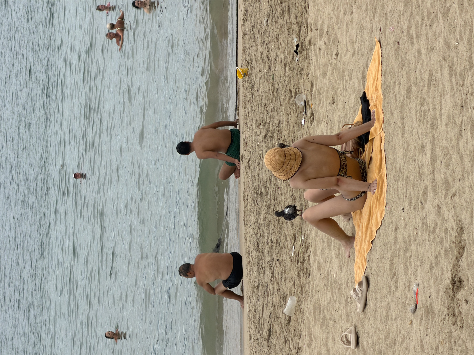 The image depicts a crowded beach scene in Nha Trang, Vietnam. In the foreground, a woman wearing a straw hat and a leopard-print bikini sits on an orange towel, facing away from the camera. Nearby, a pigeon pecks at the sand, and a pair of beige sandals rests on the beach. Further back, two men sit on the sand near the water's edge, both looking out at the sea. The water is populated with several people swimming and wading. In the background, the ocean stretches to the horizon under a cloudy sky. The beach is sandy and appears to have some litter scattered on it.