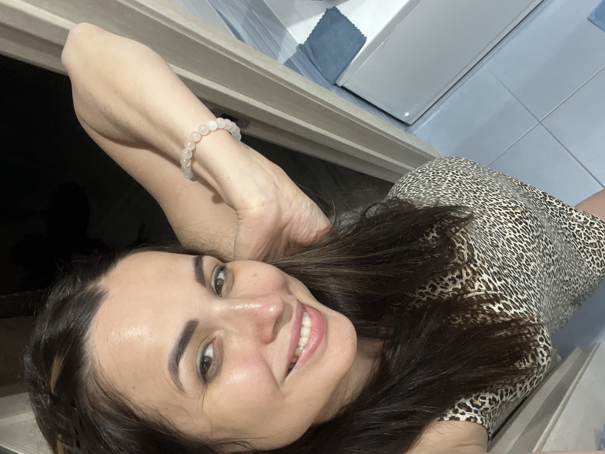 In a close-up, high-angle selfie, a woman with long, dark hair and a leopard-print top smiles. Her right arm is raised and bent, resting on a doorframe, and she wears a bracelet of light pink beads. The background includes a door, a white appliance, and pale blue tiled walls. The lighting suggests an indoor setting, possibly a bathroom or hallway.