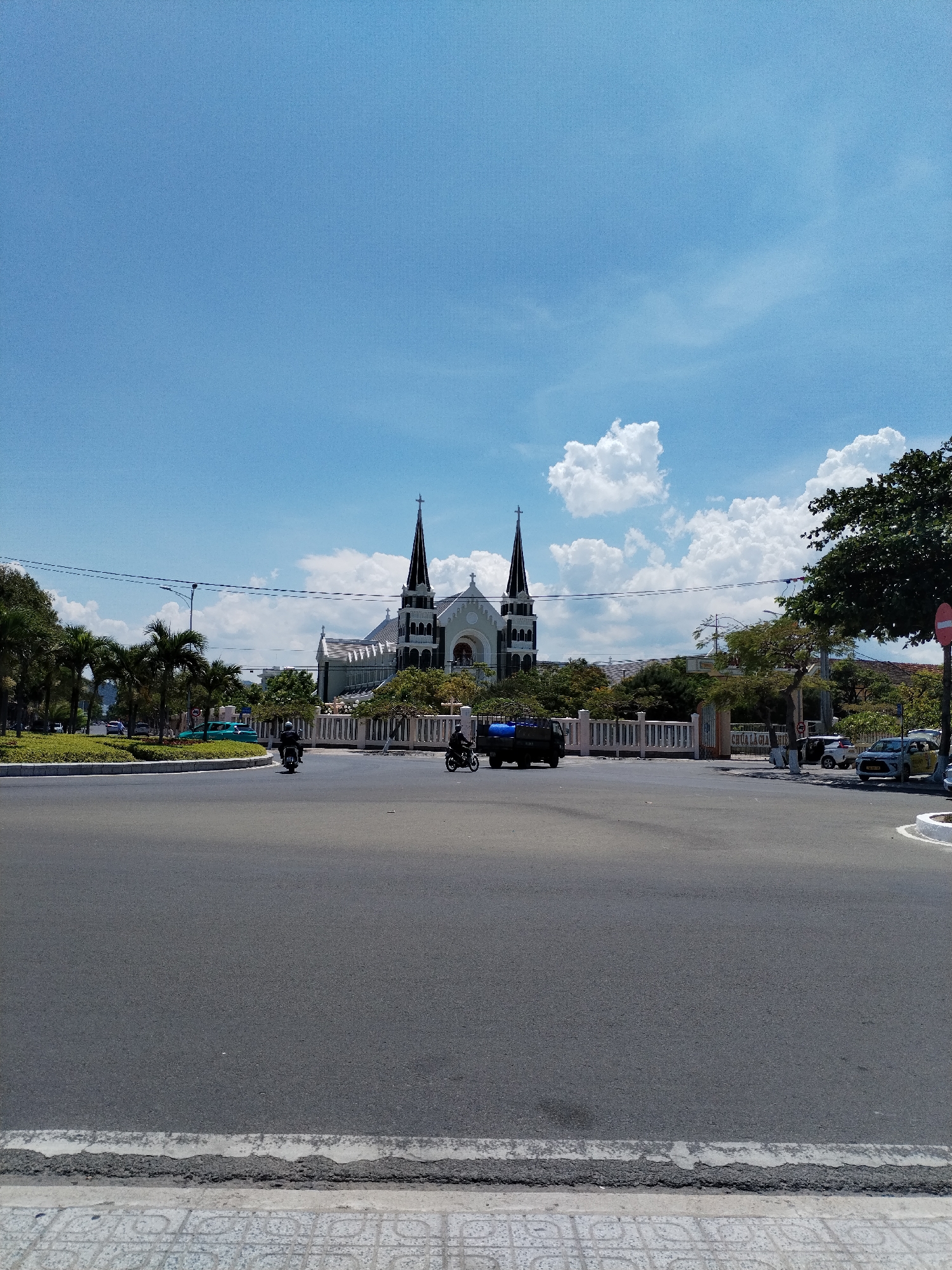 The image depicts a street scene in Bac Nha Trang Ward, Vietnam, with a church as the focal point. The church is light-colored with dark spires topped with crosses. The sky is light blue with scattered clouds. The foreground shows a paved street with two motorcycles in motion. A row of trees and shrubs lines the side of the street. Several cars are parked near the church entrance. The street is bordered by a sidewalk with a patterned edge.