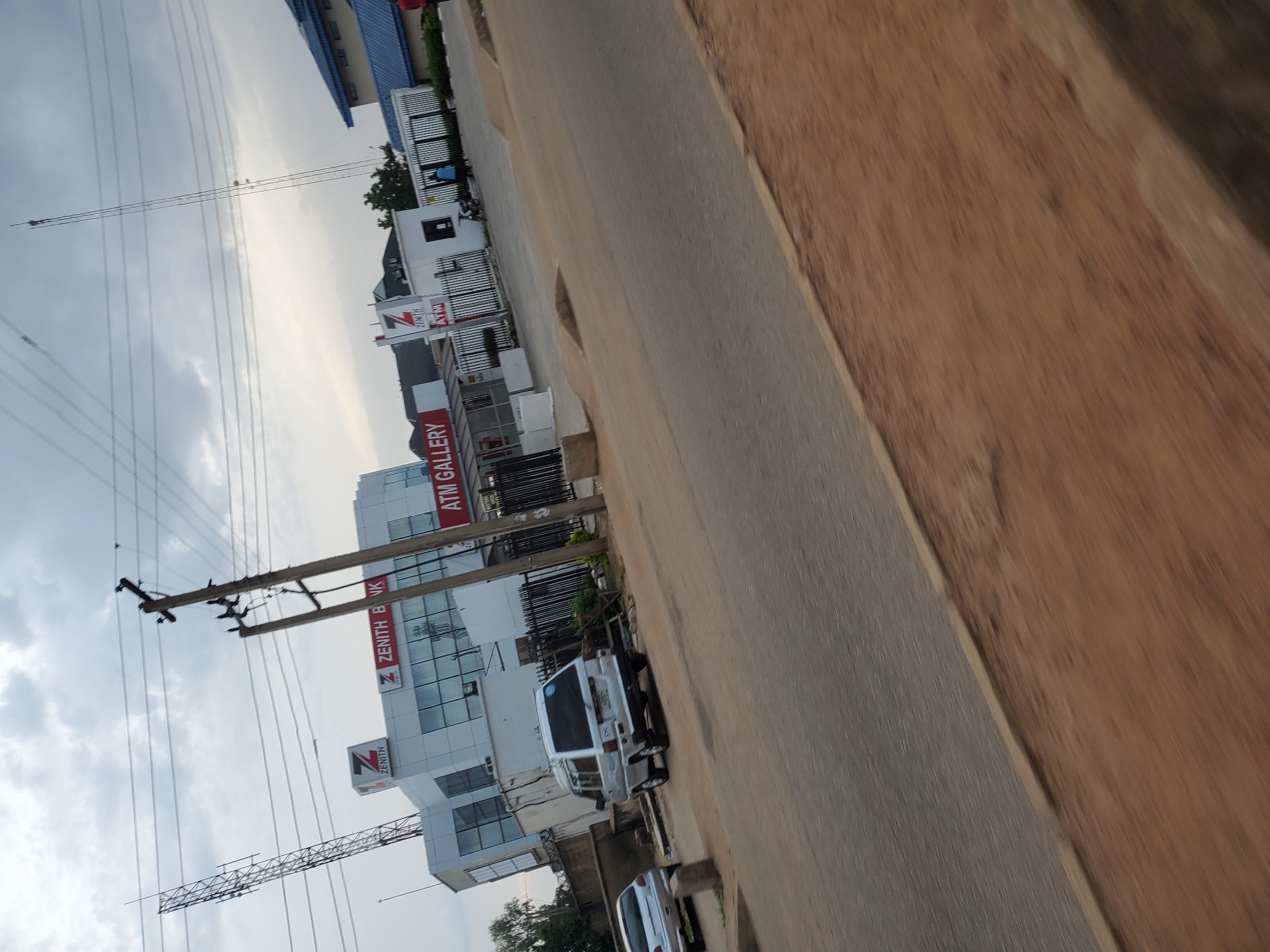 The image shows a street scene in Bukuru, Nigeria, under an overcast sky. On the left side of the image, a multi-story building with white facade and blue accents is visible. Signs on the building indicate "ZENITH BANK" and "ATM GALLERY". A white van is parked on the side of the road in front of the building. A silver car is also parked further down the street. Power lines and utility poles are prominent features of the scene, crossing the sky. The road is paved and appears to be a typical urban street. The overall atmosphere is subdued, likely due to the cloudy weather. The image is taken from a slightly elevated perspective, looking down the street.