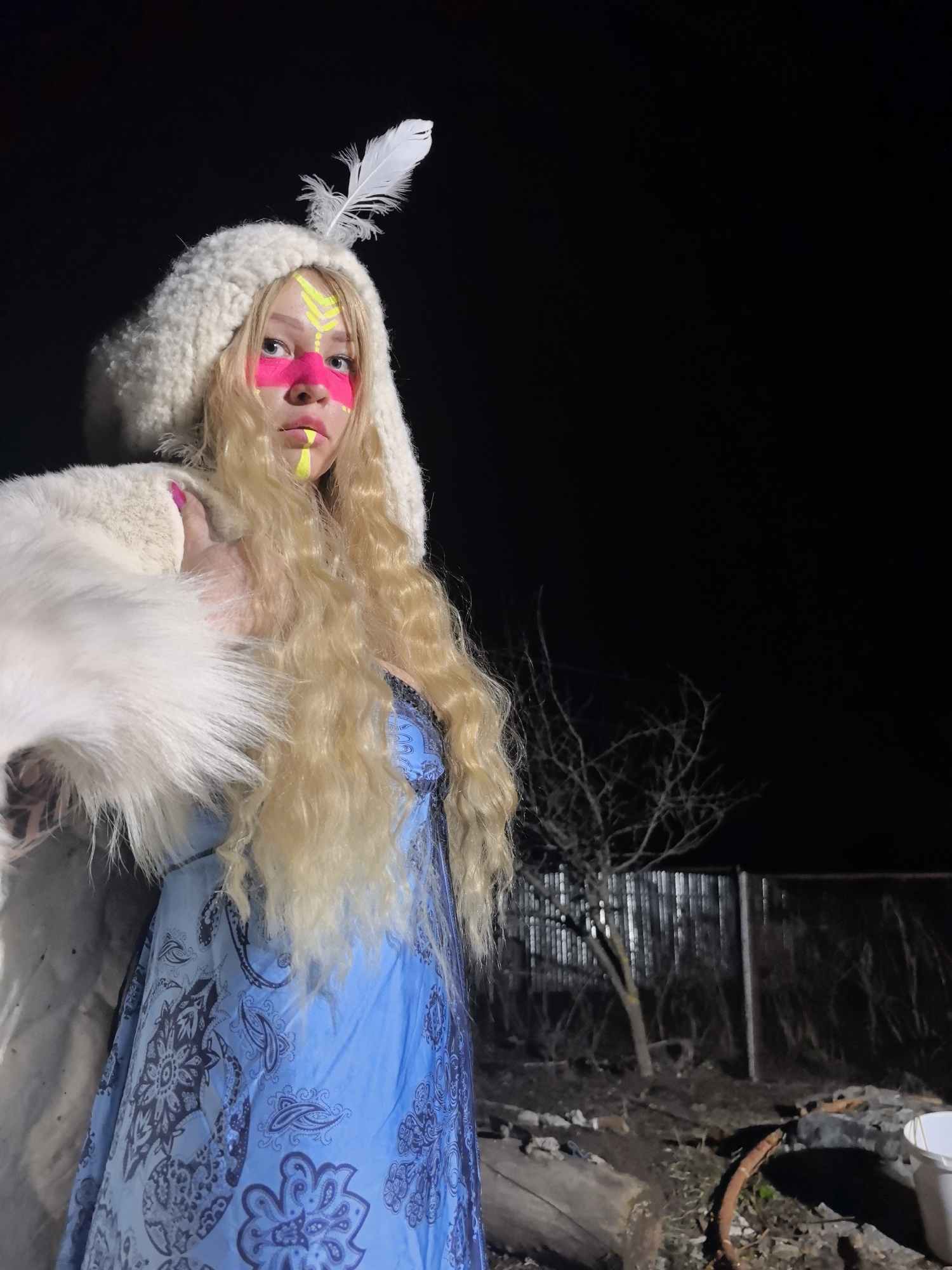 A young woman is depicted in a nighttime outdoor setting, illuminated by an artificial light source. She has long, wavy blonde hair and a distinct facial painting featuring a broad pink stripe across her eyes and nose, complemented by neon yellow patterns above and below. She is wearing a fluffy, white, hooded top adorned with a prominent white feather on the hood, and a blue dress with dark blue paisley or floral patterns. Her right hand, with pink-painted fingernails, is visible near her shoulder, with her thumb slightly raised.
The background is dark, indicating night, with a bare, leafless tree and a corrugated metal fence partially visible. The ground below is uneven, covered with logs, debris, and sparse vegetation, suggesting a rustic or semi-rural outdoor area, possibly a backyard, in Noviy, Russia. The woman's expression is neutral as she looks directly at the camera. There is no visible text in the image.