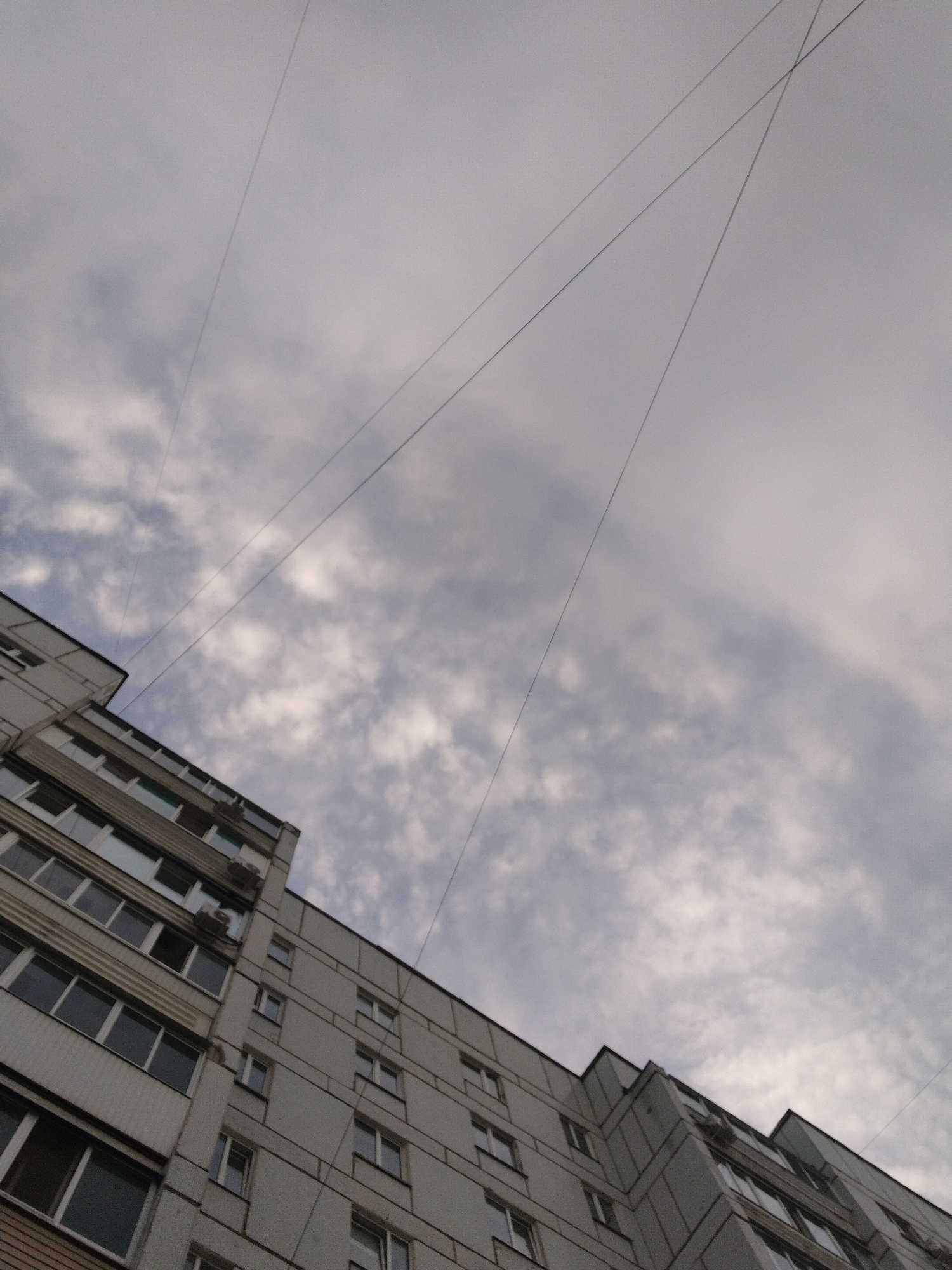 The image depicts a multi-story residential building, likely an apartment complex, under a cloudy sky. The building is constructed of concrete panels and features rows of rectangular windows. Some windows have air conditioning units installed. Overhead power lines are visible against the sky. The location is urban, but the specific city and country are unknown. There are no people visible in the frame. The overall scene appears to be a typical urban environment.