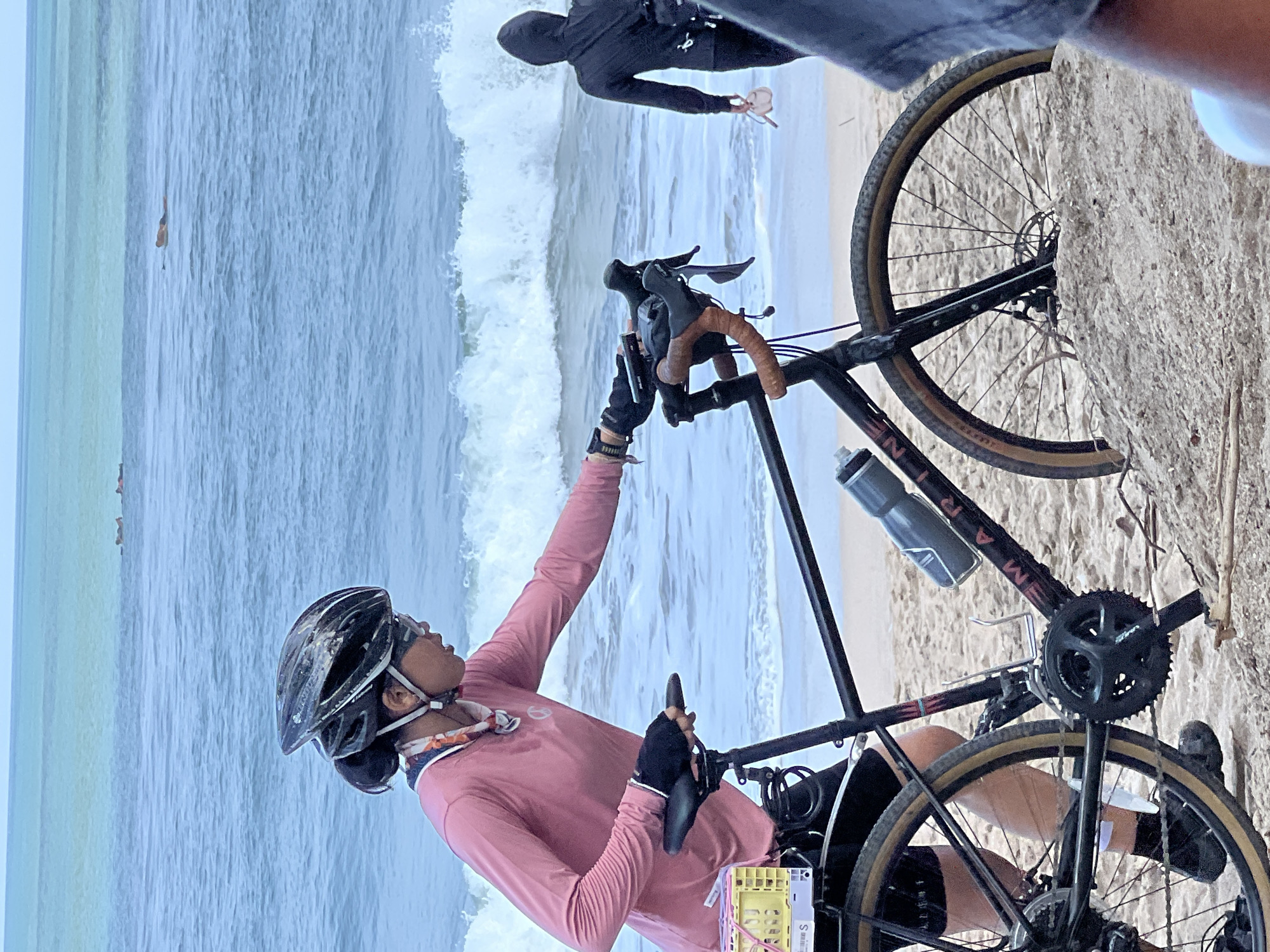 In Kuta, Indonesia, a person wearing a pink long-sleeved shirt, black helmet, and sunglasses is seen next to a bicycle on a sandy beach. The person is reaching out towards the bicycle's handlebars. The bicycle is black with brown handlebars and has a water bottle attached to the frame. In the background, the ocean waves are breaking on the shore, and in the distance, several people are visible in the water, some appearing to be lying on surfboards. Another person wearing a black hooded jacket is walking away from the viewer towards the ocean. The scene depicts a coastal environment with activities suggesting outdoor recreation.