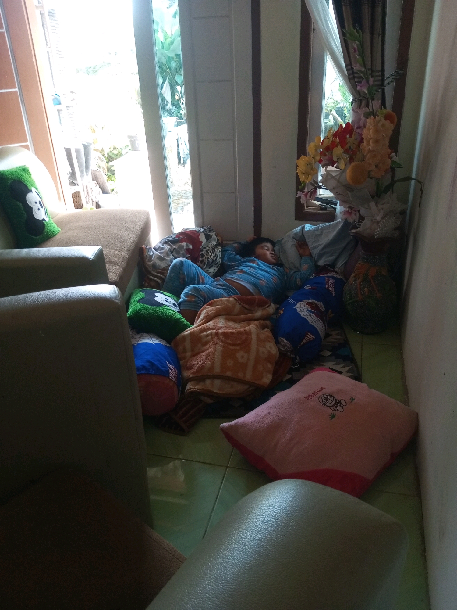 A young child is asleep on the floor, surrounded by numerous pillows and blankets. The child is wearing blue pajamas. The scene appears to be indoors, possibly in a living room or bedroom. In the background, there is a window with curtains and a vase of flowers. A sofa is partially visible in the foreground. The flooring is tiled and appears to be green. The overall lighting suggests it is daytime. The location of Krasak, Indonesia, is not discernible from the visual information provided.