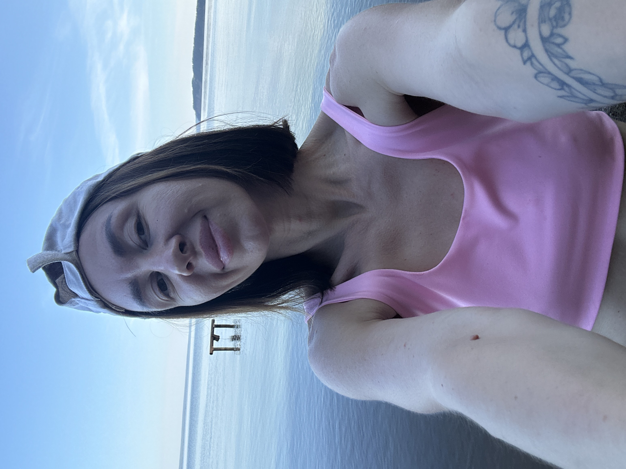 A woman is taking a selfie on the shore of a body of water, possibly a lake or river, in the city of Galovo, Russia. The sky is a clear blue with a few wispy clouds. In the distance, there is a small wooden structure partially submerged in the water, and a distant shoreline with trees. The woman is wearing a pink tank top and a light-colored baseball cap. Her short, dark hair is visible beneath the cap. Her left arm is extended forward, holding the camera, and a tattoo of a floral design is visible on her right arm. The water is calm and reflects the blue of the sky. The overall scene suggests a casual outdoor setting.