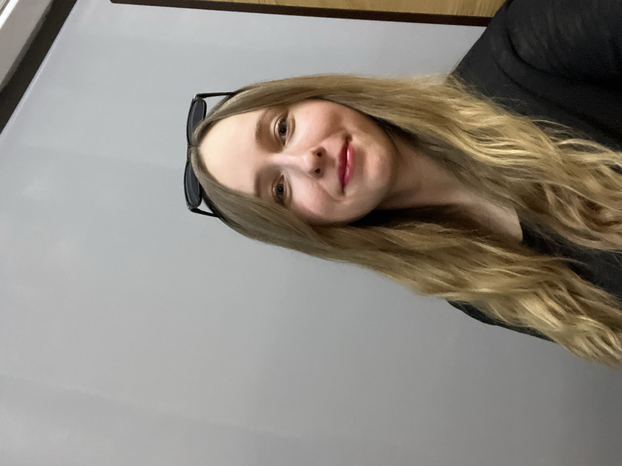 A blonde-haired woman is posing for a selfie. She is wearing sunglasses on her head and a black top. The background is a grey wall. The location is not identifiable as Mytishchi, Russia from the provided image.