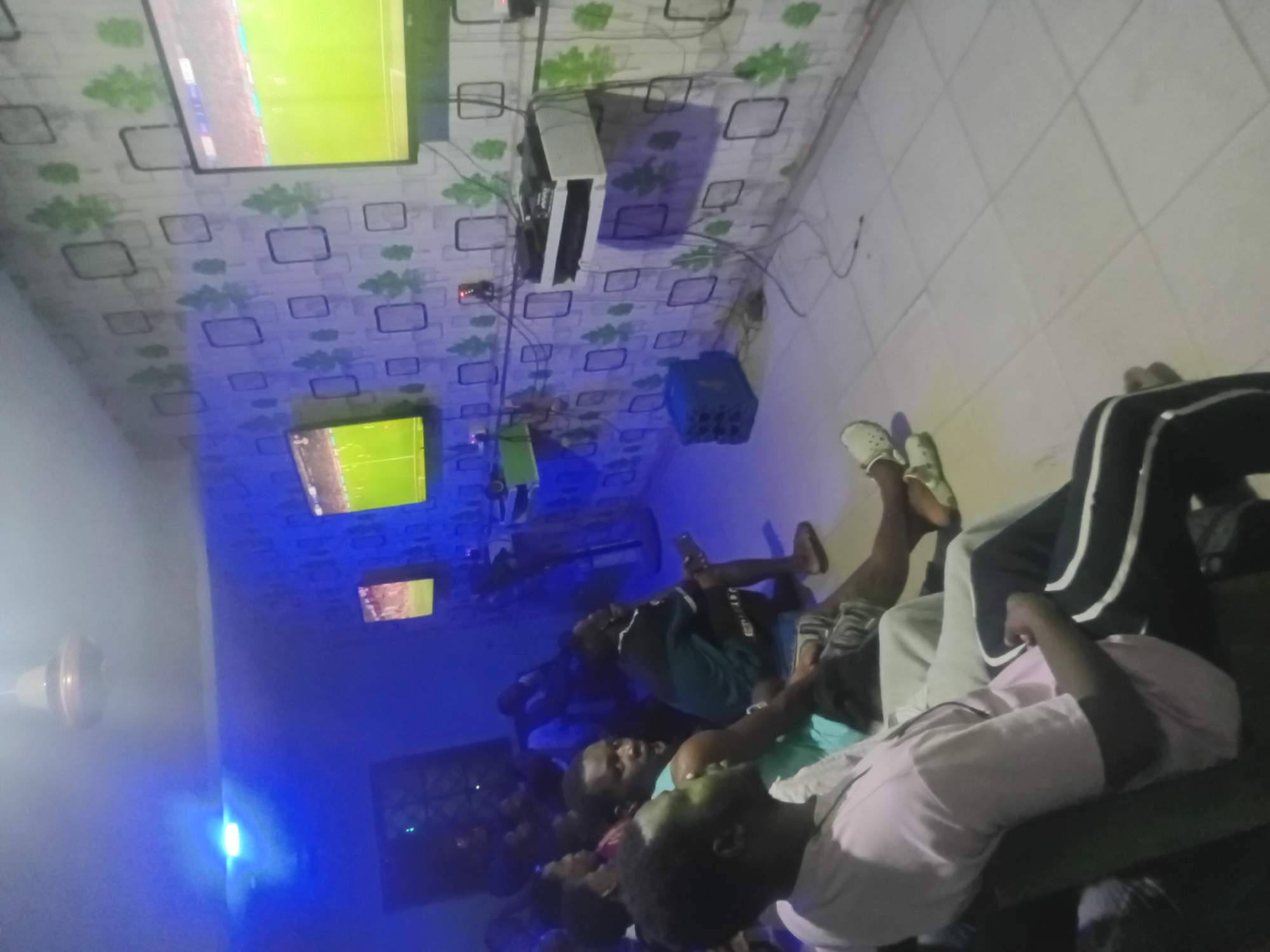 A low-angle indoor shot depicts a group of individuals gathered in an unknown location, city unknown. The room is illuminated by blue light and features multiple televisions mounted on the walls, all displaying what appears to be a soccer match. The walls have a white square with green floral pattern. The floor is tiled in white squares. The individuals are mostly seated on what seems to be a couch, their gazes directed at the TVs. Some are wearing casual clothing. Wires and an air conditioner are visible on the upper portion of the wall.