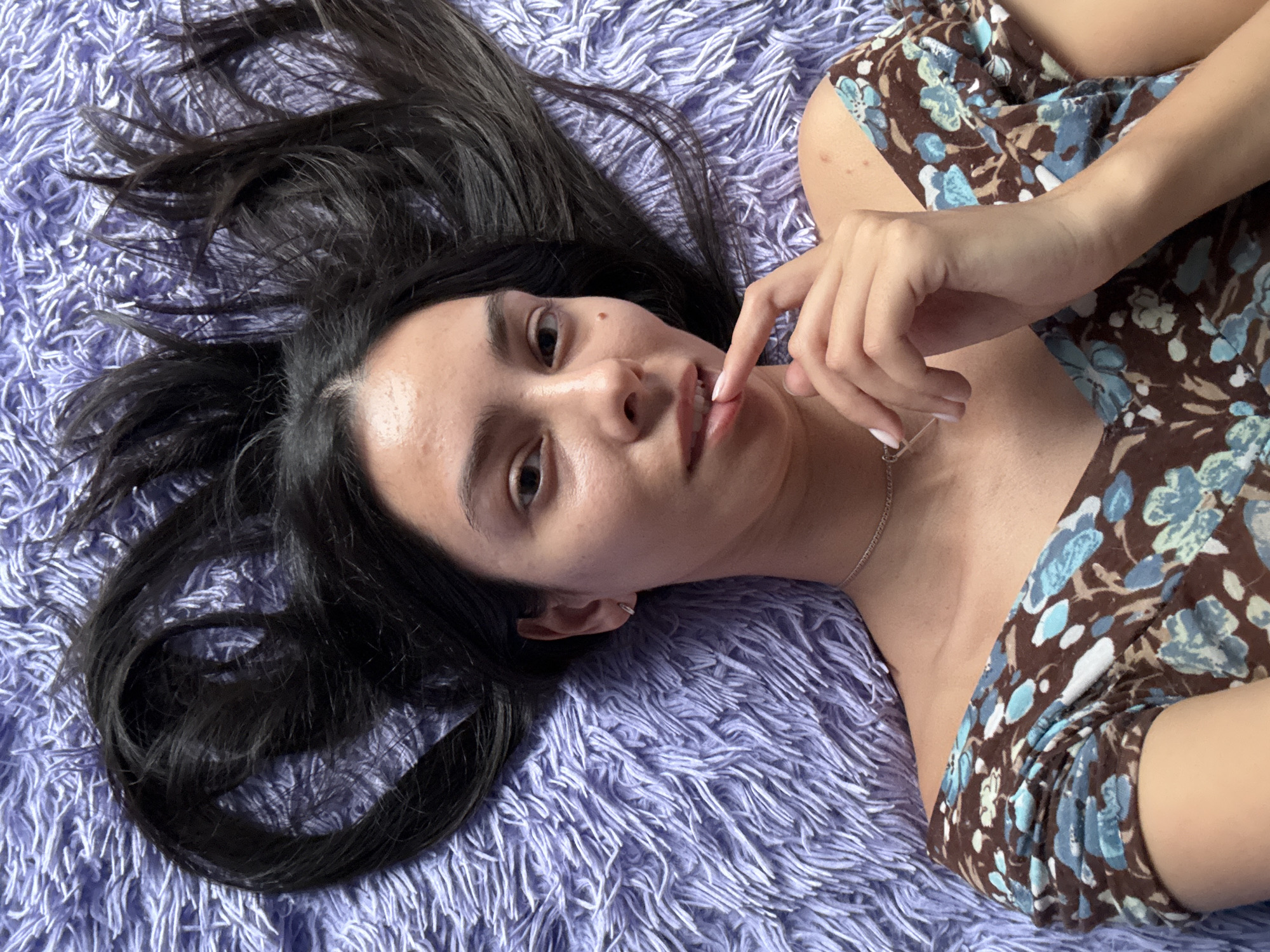 A young woman lies on a purple, shag rug. She has long, dark hair spread around her head on the rug. She is wearing a brown and blue floral dress. Her right hand is raised with her index finger touching her lips. She is wearing a silver necklace. The location is not specified in the image. There is no indication that the location is in the city of Chaikovsky, Russia.