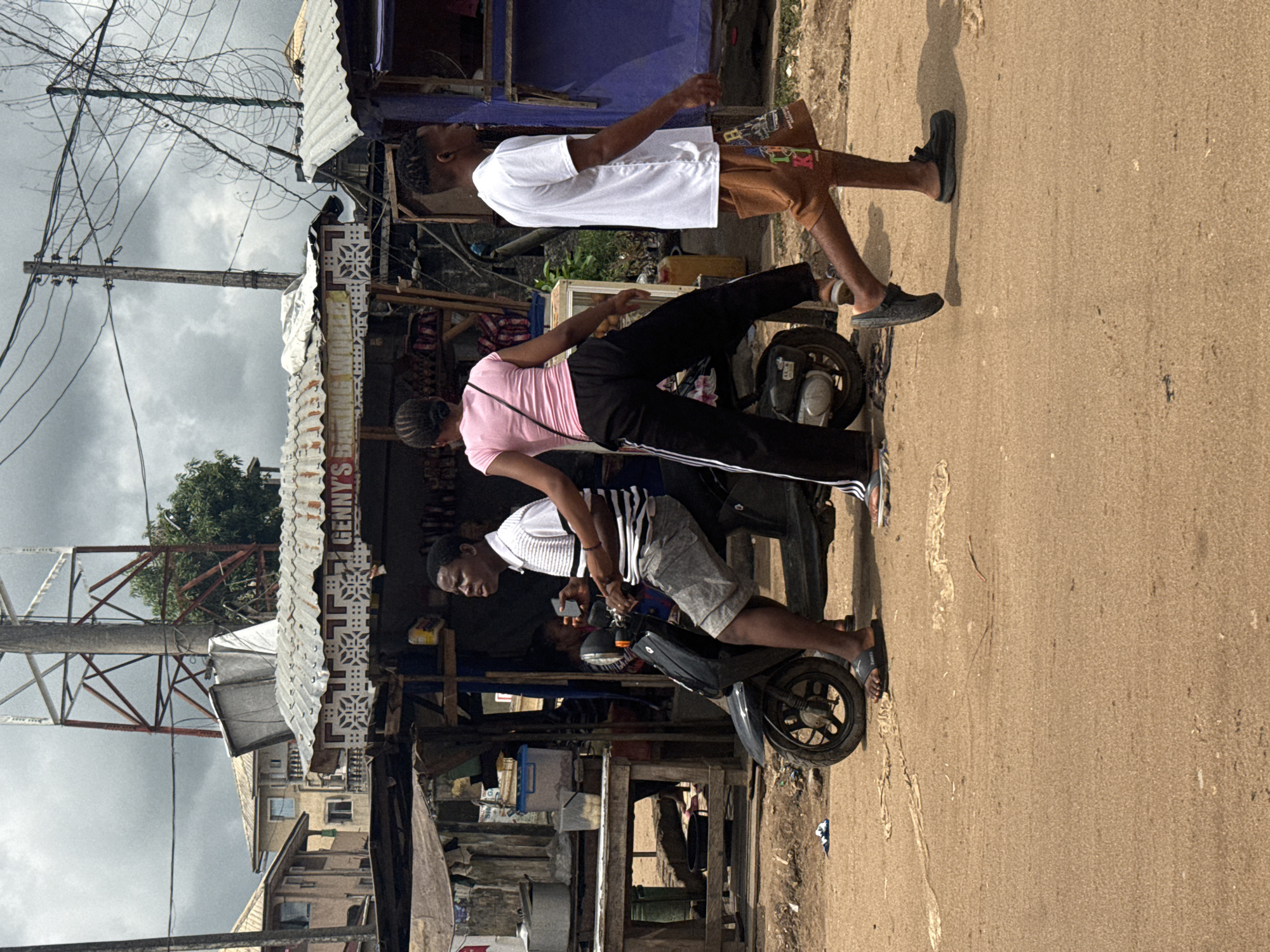 This media file captures a bustling street scene in Ihiagwa, Nigeria, under an overcast sky during daytime. The image features three individuals on a paved road, flanked by informal market stalls and structures.
In the foreground, a man wearing a white and black striped polo shirt and grey shorts is seated on a black scooter, looking down at a mobile phone in his hands. Next to the scooter, a woman in a pink t-shirt and black track pants with white stripes stands, facing away from the viewer, appearing to wait or observe. Further right, another man, dressed in a white t-shirt and brown patterned shorts, stands on the roadside, looking towards the left side of the frame. Distinct shadows are cast on the road, indicating daylight.
The background reveals corrugated metal roofs of market stalls and various buildings. Overhead, numerous entangled electrical wires crisscross, connected to a wooden utility pole. A visible sign on one of the market stalls reads "GENNY'S". The scene depicts typical daily street activity in a local community setting.